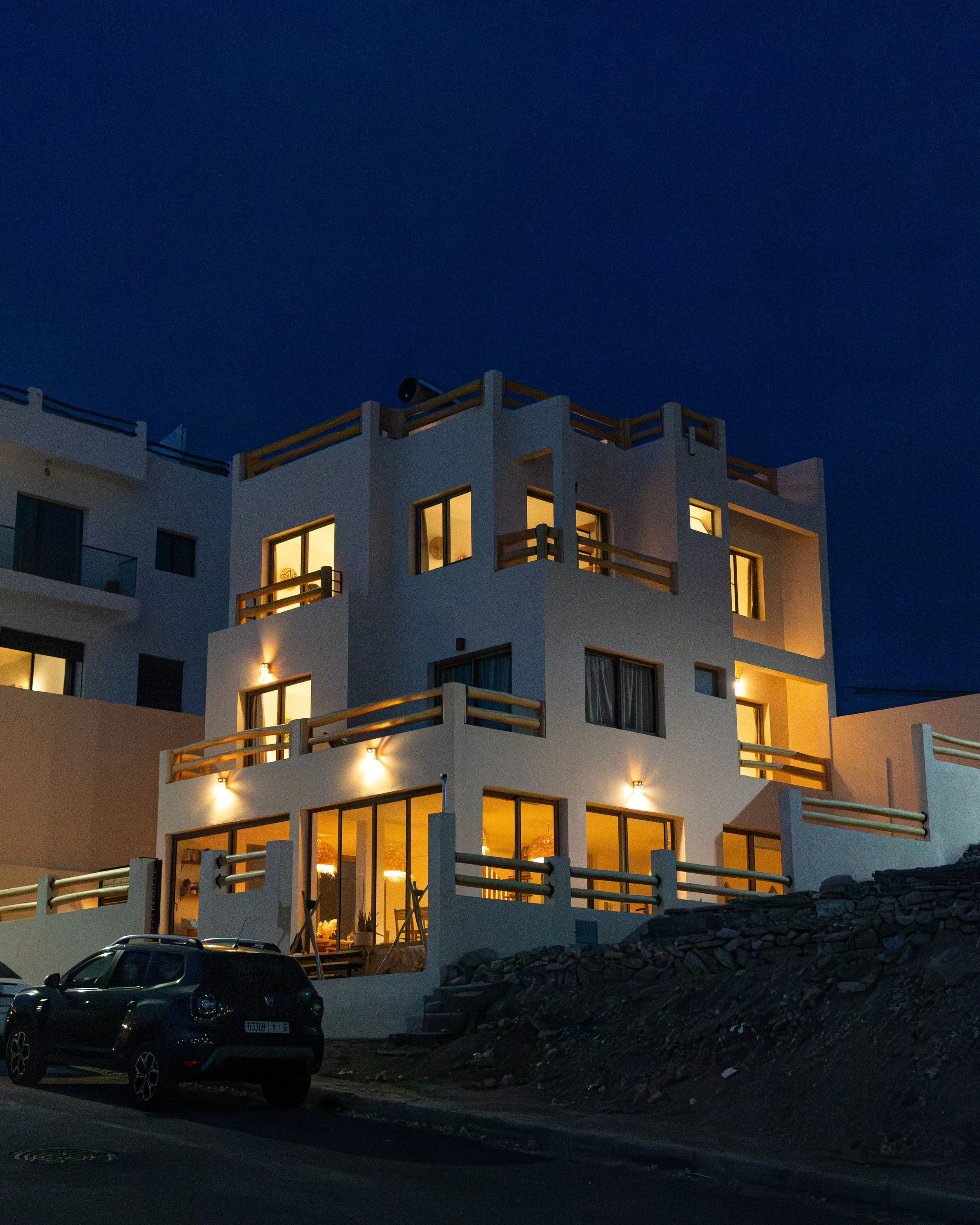Un immeuble moderne blanc avec plusieurs grandes fenêtres lumineuses à l'intérieur, situé dans un environnement sombre. Un véhicule noir est stationné devant l'immeuble, et le ciel est entièrement nuit. Gros travaux de construction sur un terrain rocheux en face de l'immeuble.