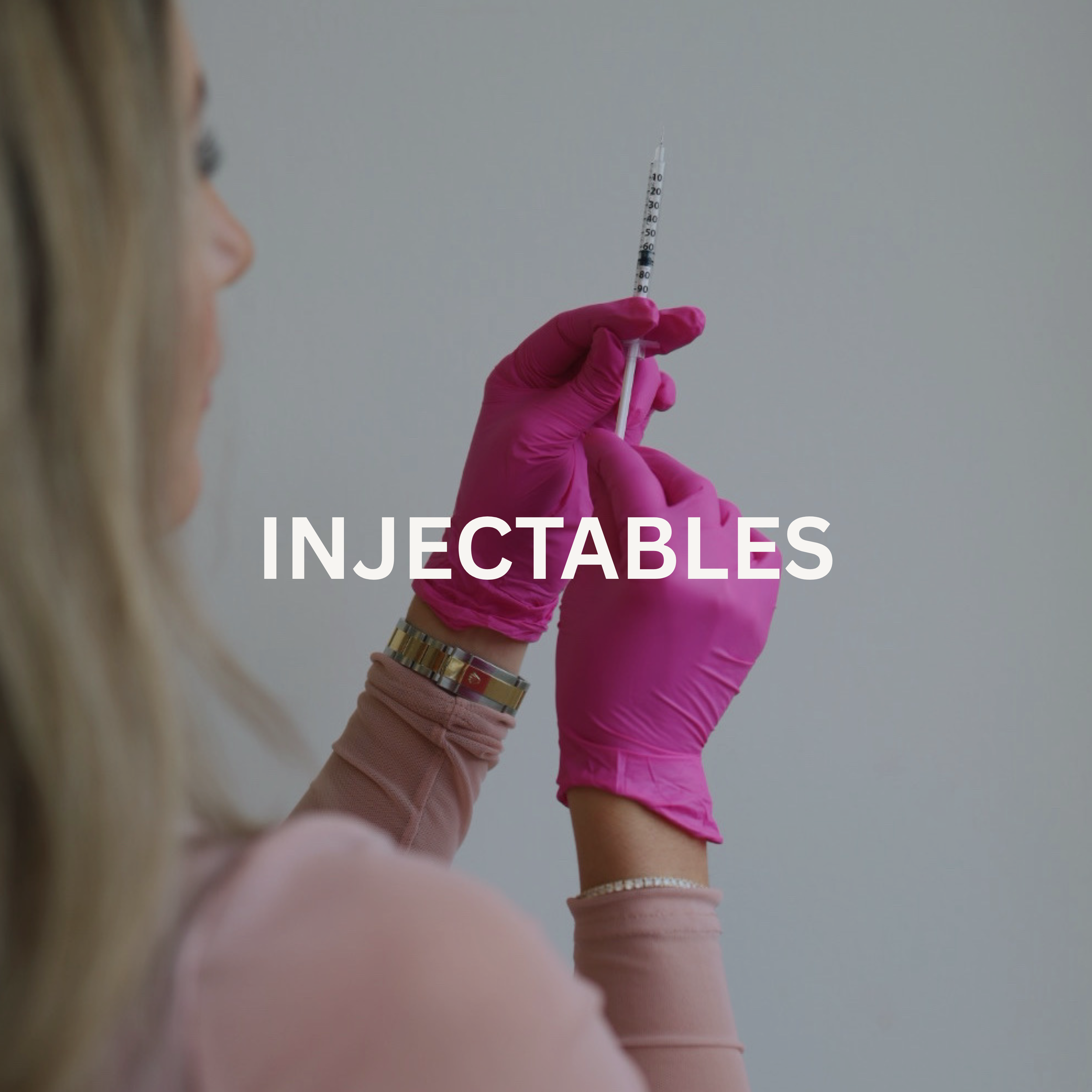 A healthcare professional wearing pink gloves holds a syringe filled with a clear liquid, preparing to administer an injection. The focus is on the syringe, with the person's face and surroundings blurred out.