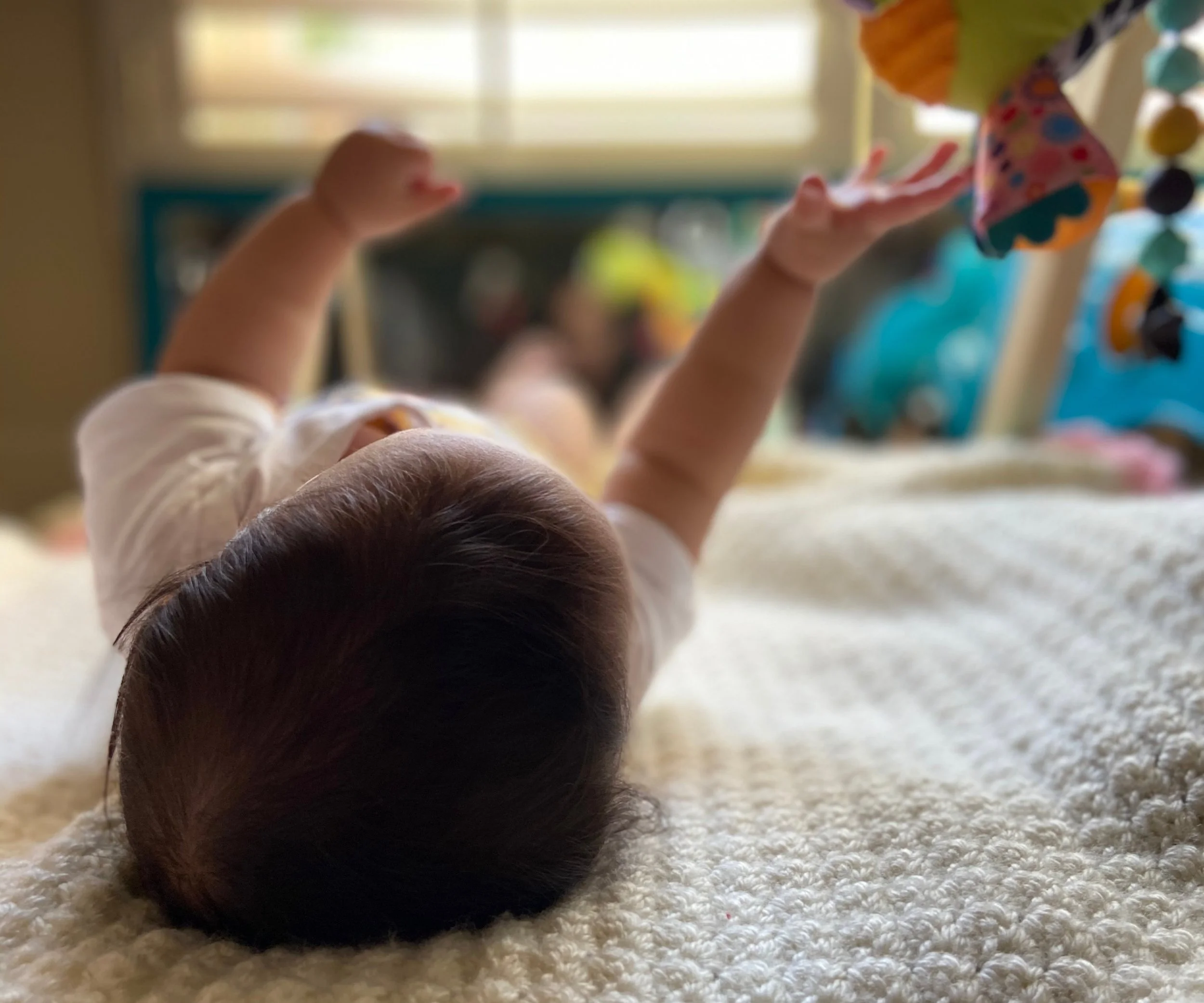 A child's head and arms on the carpet as they reach out towards colorful objects hanging above, with a window in the background at an occupational therapy session with Jacklynn Knotts, OTR/L, IBCLC at Tiny Feeding Co. in El Paso, TX.