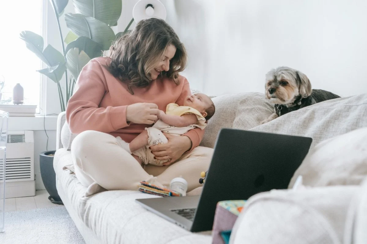 A woman holding a baby on a couch with a dog sitting nearby and a laptop is open in front of them on a virtual consultation with Jacklynn Knotts, OTR/L, IBCLC at Tiny Feeding Co. in the United States