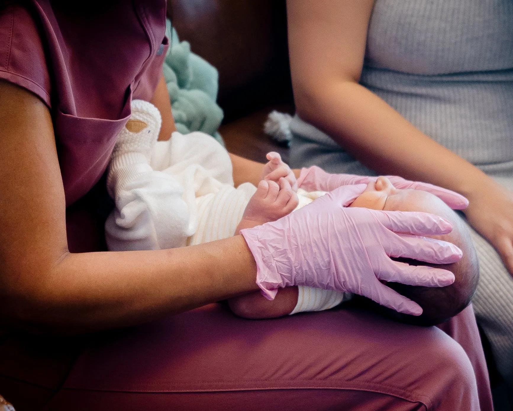 Jacklynn is holding a baby in her lap with her hands in pink gloves on either side of the baby's face to improve oralmotor skills for tongue and lip ties, breast or bottle feeding challenges, and more to provide occupational therapy or lactation.