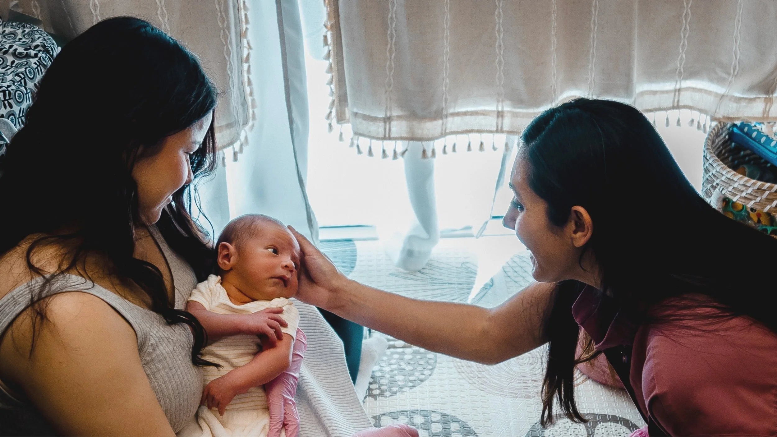 A woman holding a newborn baby on her chest with a Jacklynn Knotts, OTR/L, IBCLC in a pink shirt touching the baby's cheek inside a room with curtains and a basket in the background at Tiny Feeding Co. in El Paso, Texas
