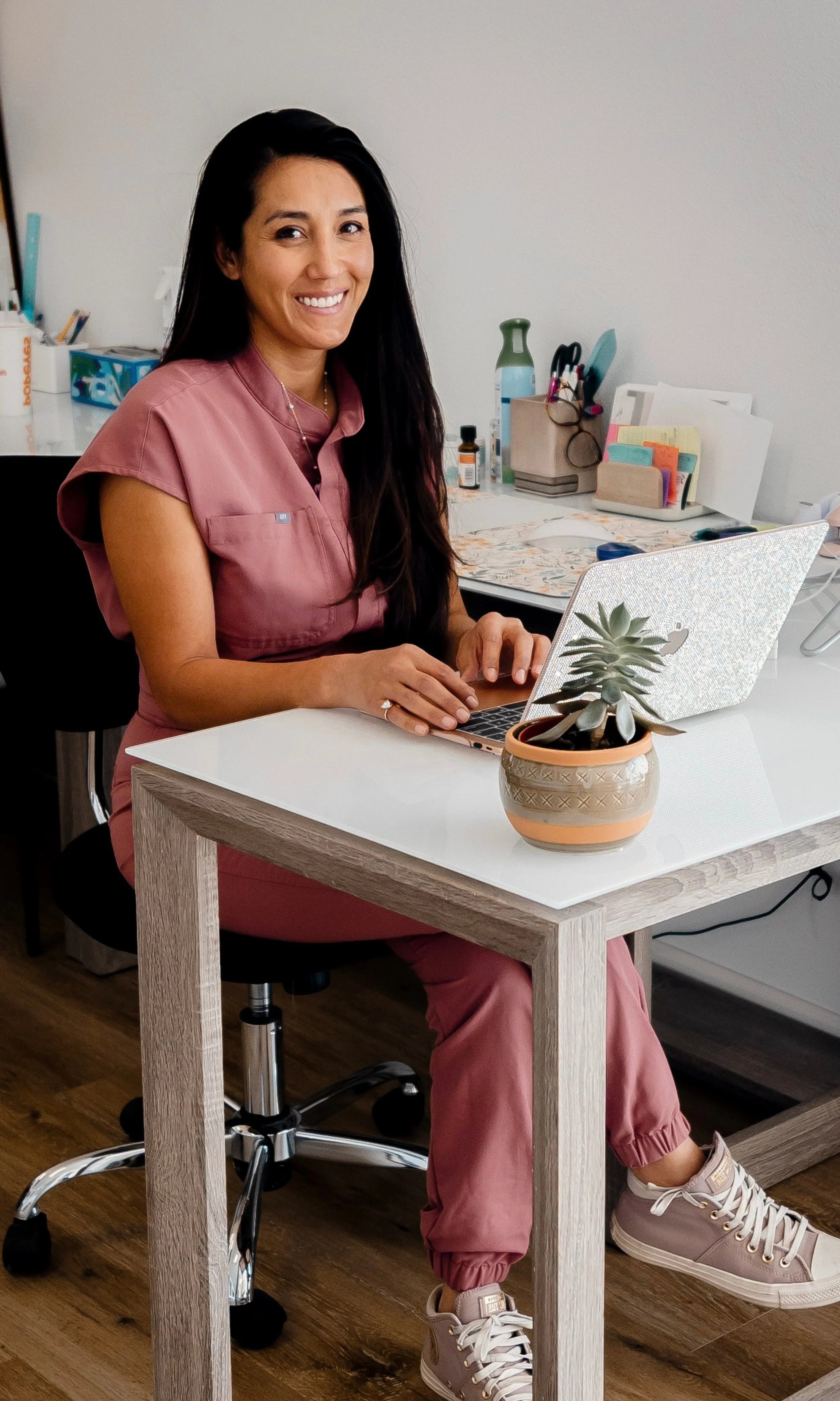 A woman with long dark hair sitting at a desk with a silver laptop, smiling. She is wearing a pink scrub top and matching pink jogger pants, with beige sneakers. In front of her is a potted succulent plant. The desk has various office supplies, including a pair of glasses, a water bottle, and a small medicine bottle, with a white wall in the background.