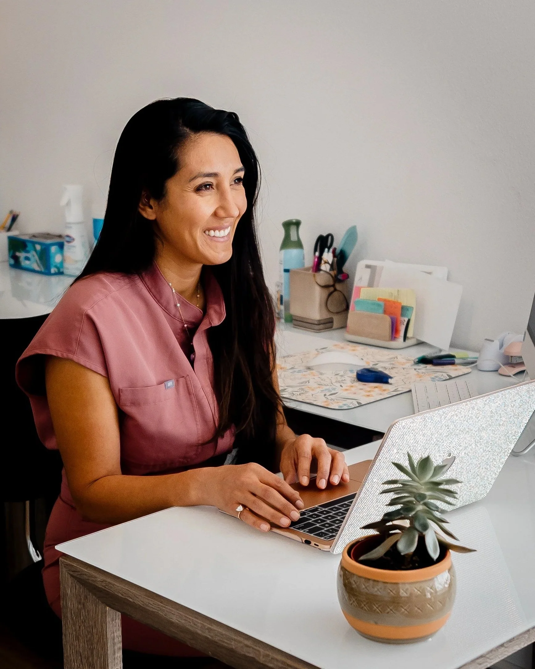 Jacklynn Knotts sits at a desk with long dark hair down one shoulder smiling off-camera. She is wearing pink scrubs and sitting in an office with a plant in the front. Jacklynn owns Tiny Feeding Co., a lactation & OT clinic in El Paso, Texas