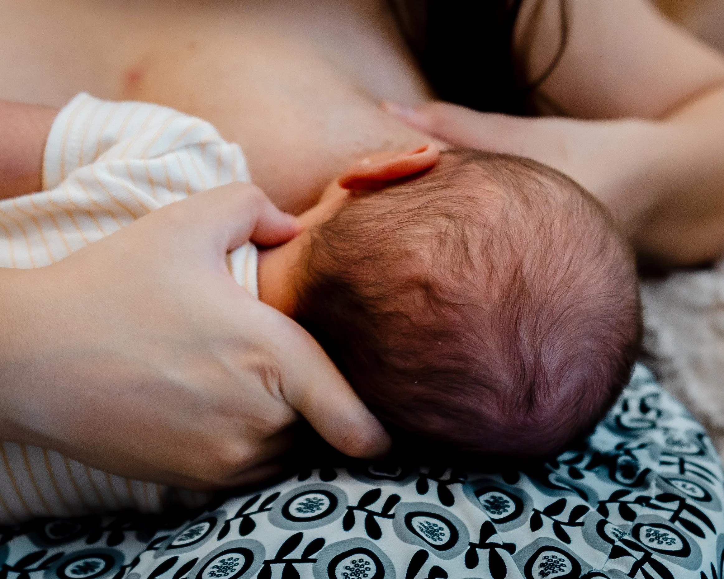 Close-up of a newborn baby nursing from a mother's breast, with the mother's hand gently holding the baby's head, and the baby lying on a patterned surface.
