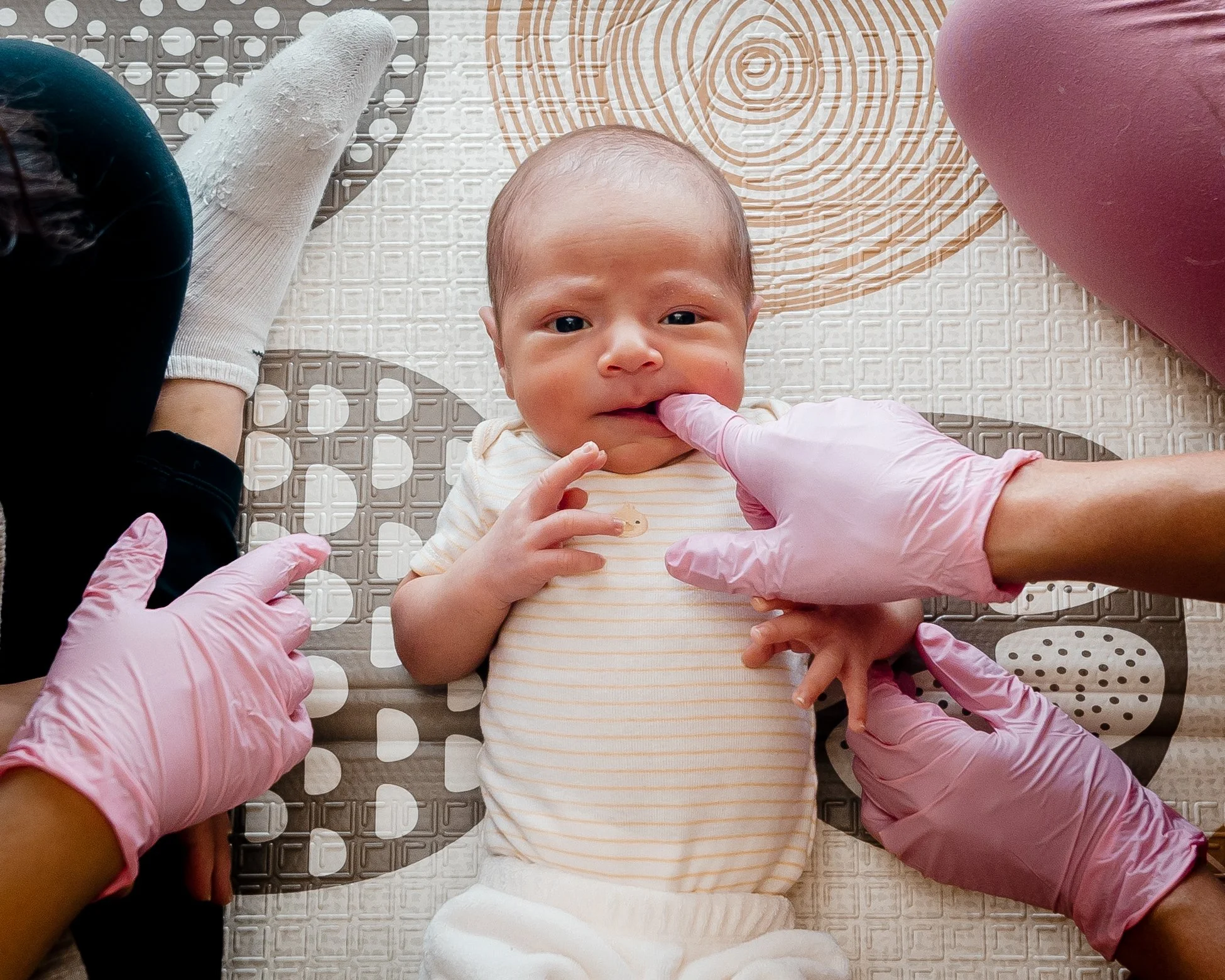 A baby lying on a patterned mat while the OT wearing pink gloves examine the baby's mouth and chest Pink scalloped edge with a transparent background at Tiny Feeding Co. in El Paso, Texas where they work with moms and babies for feeding.