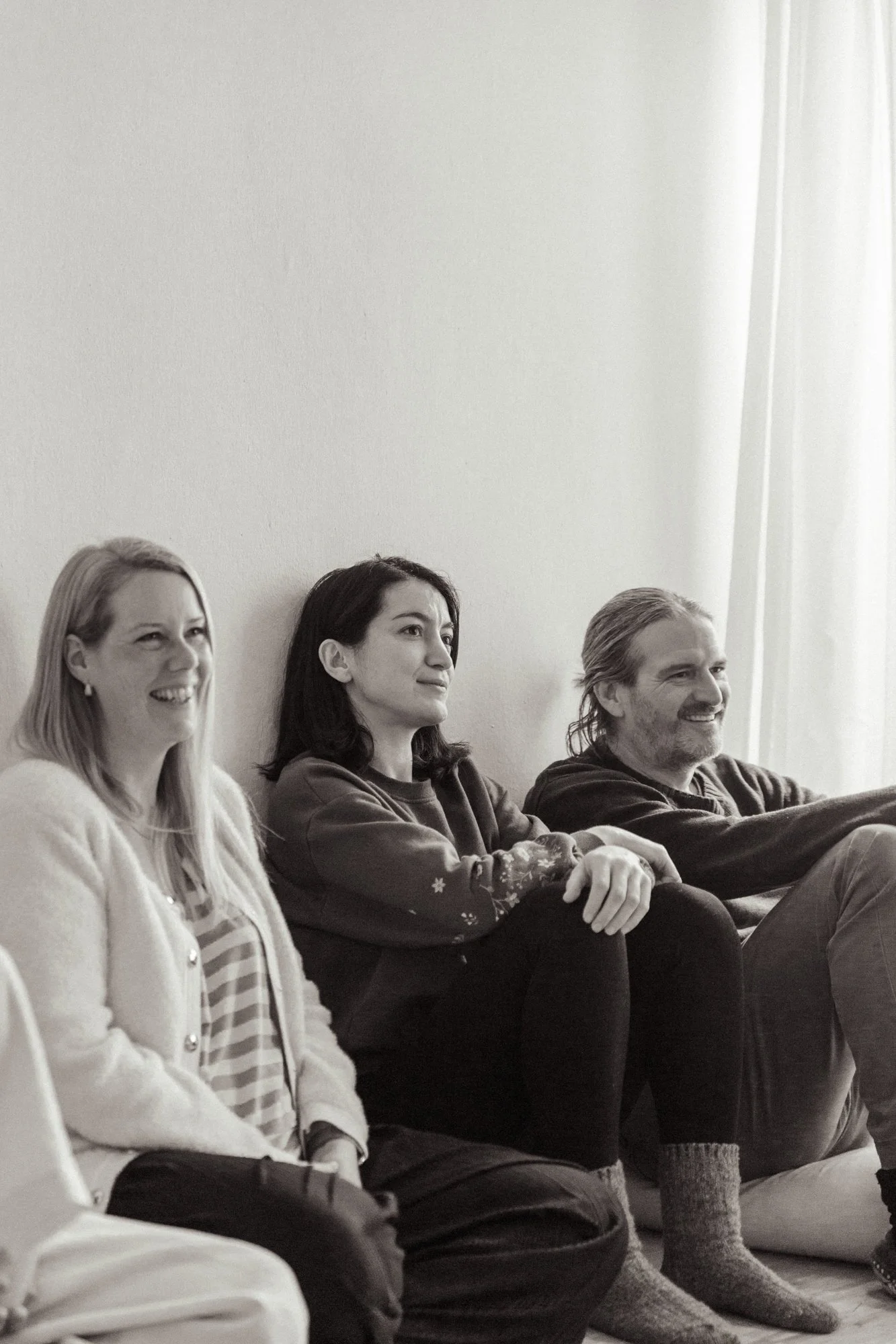 Drei Menschen sitzen entspannt auf dem Boden in einer Reihe, lachen und schauen in eine Richtung, vor einem weißen Wand mit Vorhängen auf der rechten Seite.