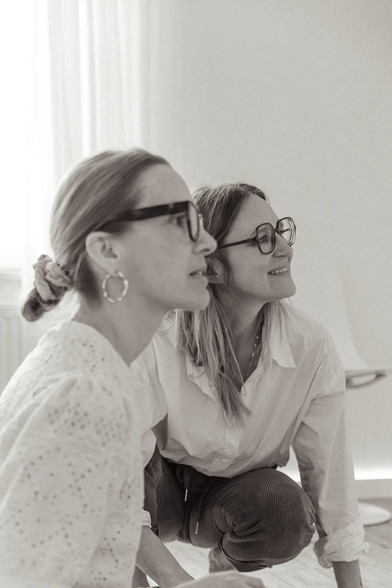Zwei Frauen mit Brillen, eine lacht, die andere schaut interessiert, sitzen auf dem Boden in einem Raum mit natürlichem Licht.