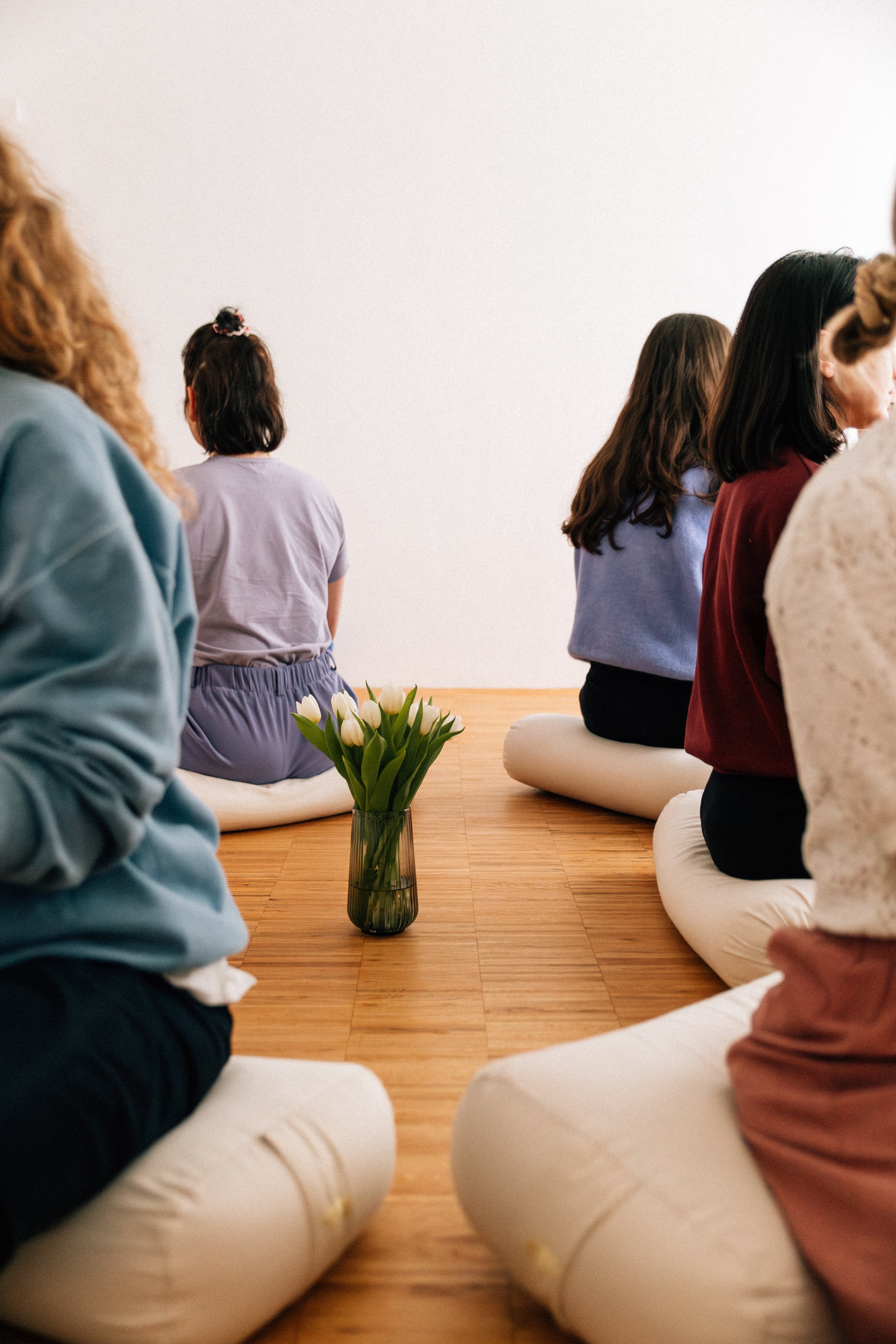 Menschen sitzen in Schneidersitz auf Kissen in einem Raum mit weißer Wand, es steht eine Vase mit weißen Tulpen in der Mitte des Bodens.