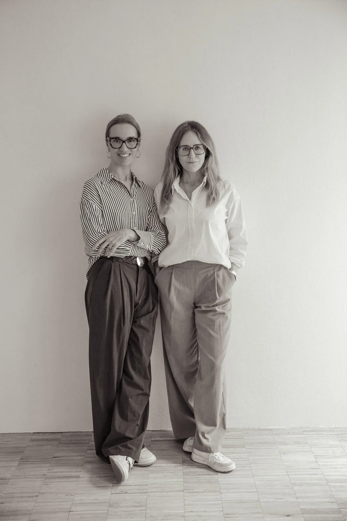 Zwei Frauen stehen nebeneinander vor einer weißen Wand. Beide tragen Brillen, eine trägt ein gestreiftes Hemd und die andere eine weiße Bluse, beide haben lockiges Haar. Die Frauen lächeln leicht und posieren entspannt.