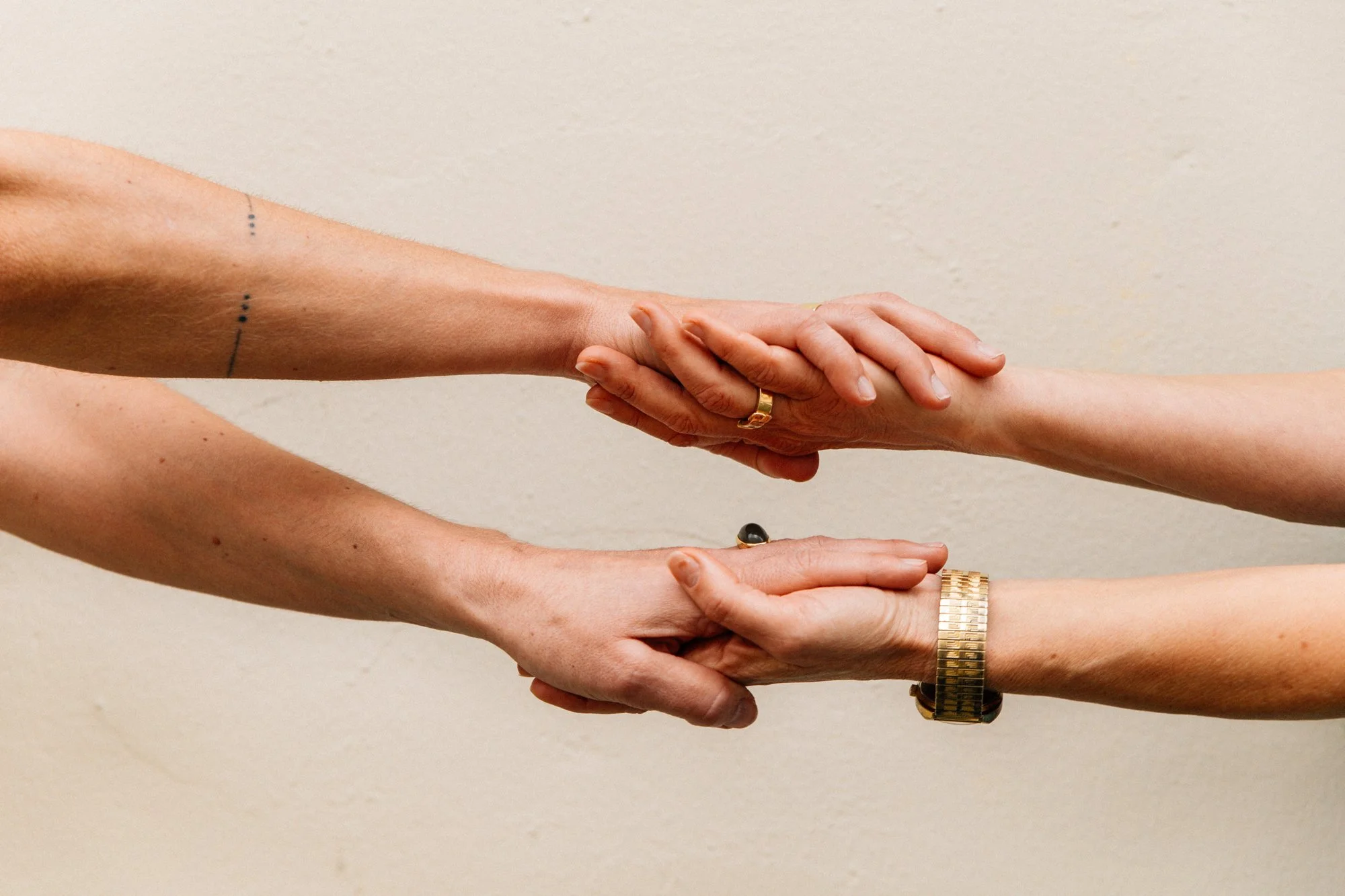 Zwei Menschen halten sich an den Händen, die obere Hand trägt eine goldene Uhr und eine Ring, während die untere Hand einen Ring mit schwarzem Stein trägt.