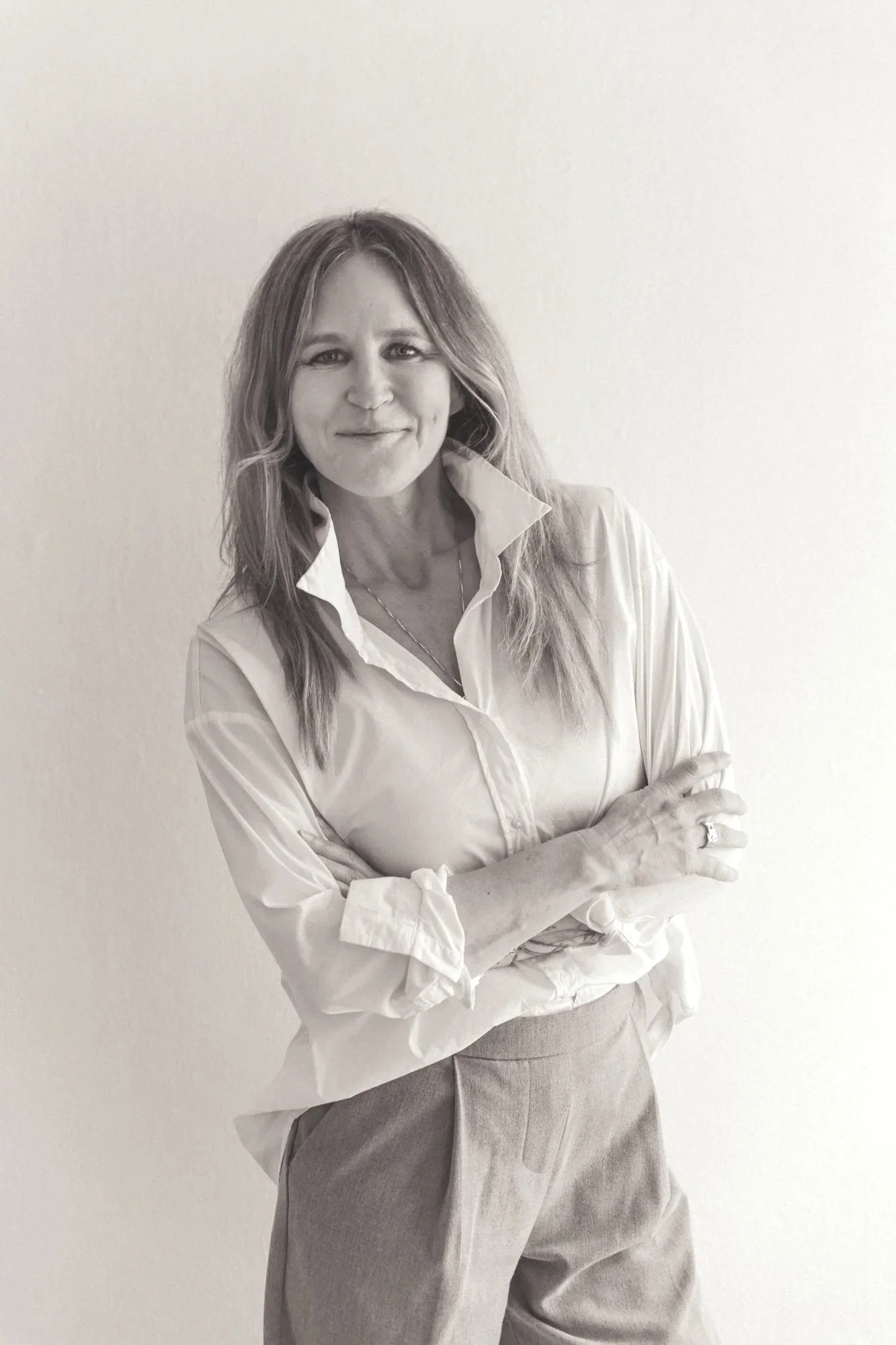 Schwarz-weiß-Fotografie einer Frau, die ein weißes Hemd trägt, die Arme verschränkt und vor einer weißen Wand steht.