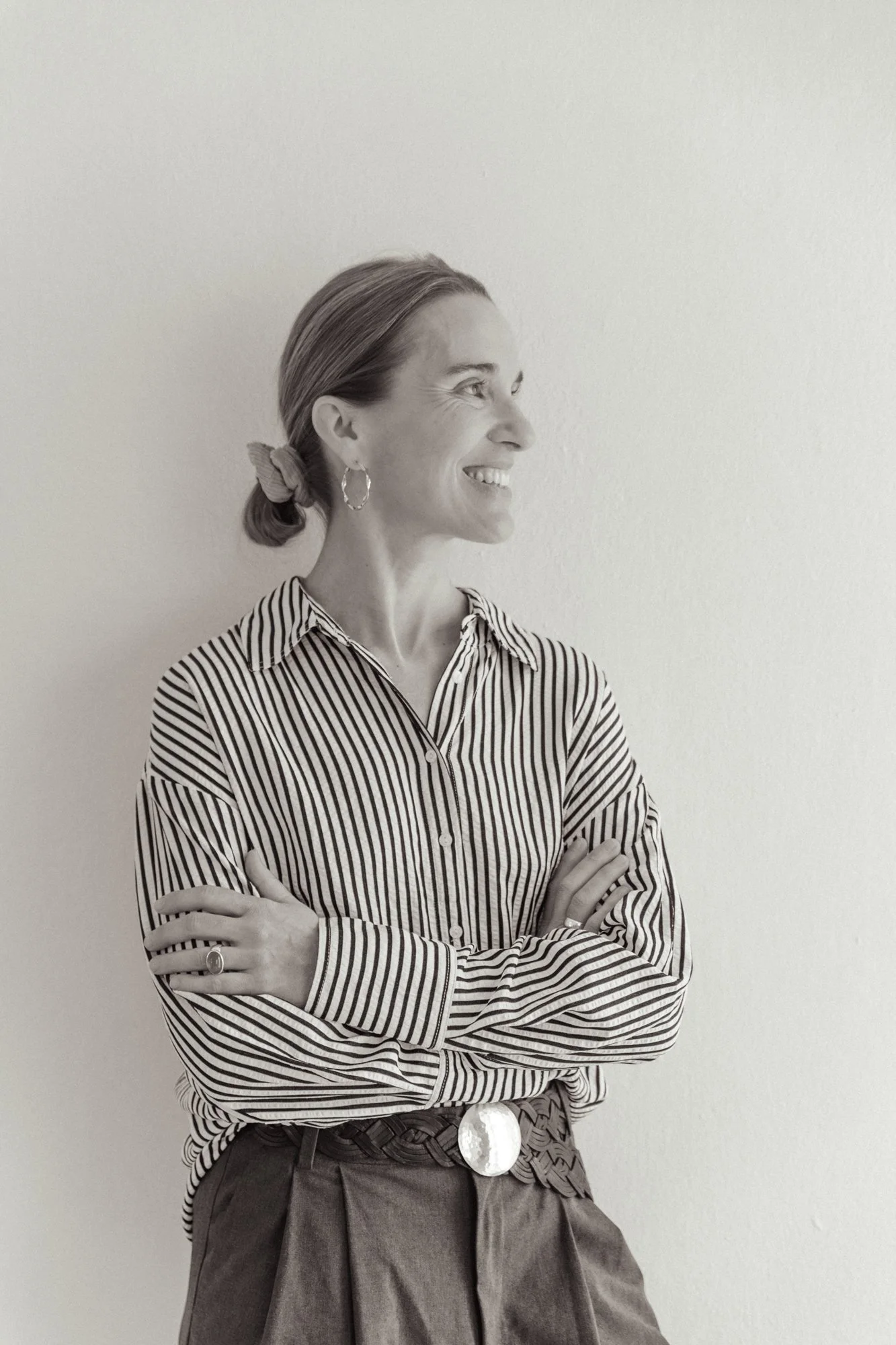 Schwarz-weiß-Fotografie eines lächelnden, lachenden Frau in einem gestreiften Hemd und mit Haarband, die seitlich gegen eine leere Wand steht.