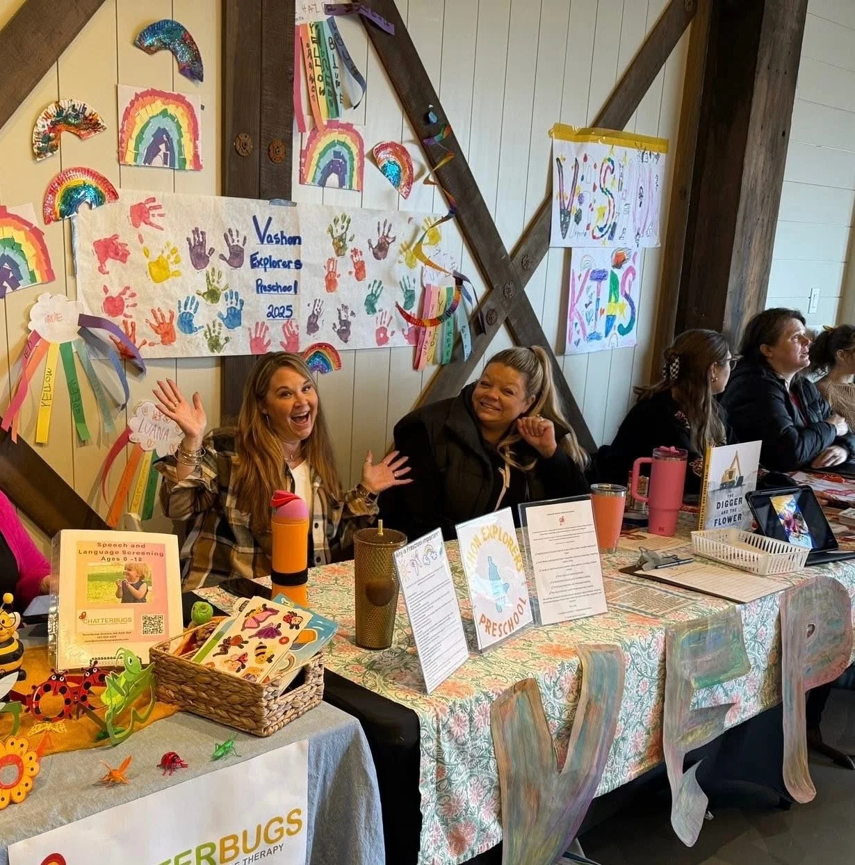 School registration or informational event table with children's artwork, colorful decorations, and smiling women seated behind the table.