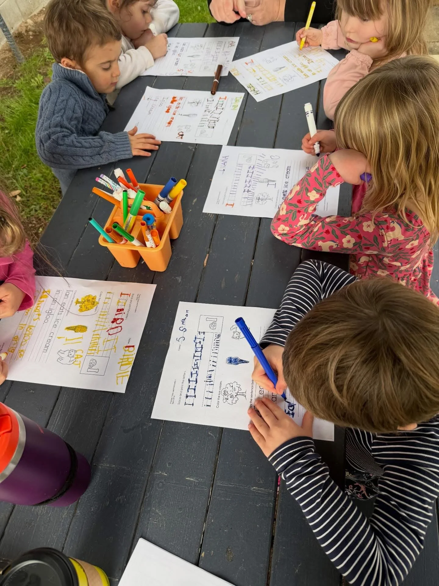 Children sitting at a black picnic table outdoors, working on colorful handwriting and drawing worksheets, with a container of markers in the middle.