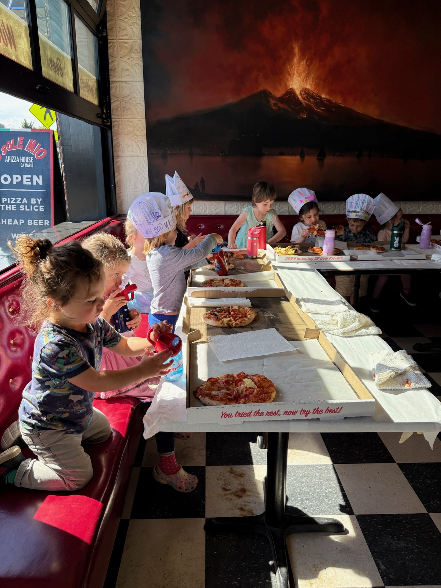 Children sitting at a long pizza restaurant table eating pizza, some wearing paper chef hats, inside a restaurant with a large volcano illustration on the wall behind them, and a sign outside advertising pizza, beer, and being open.