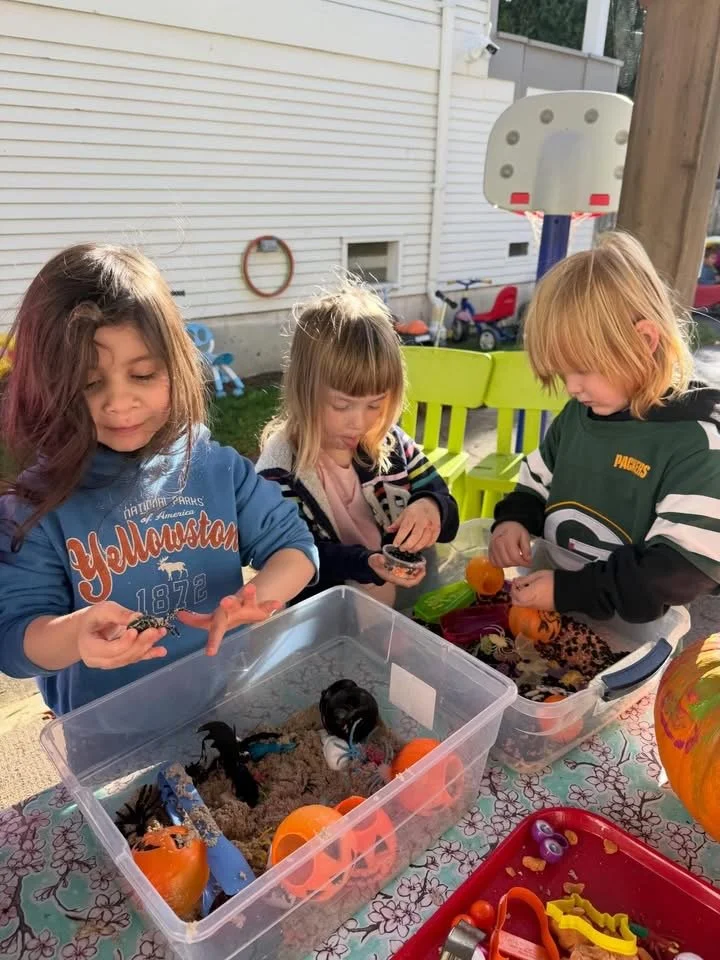 Three children playing with Halloween-themed toys outdoors at a table with a Halloween tablecloth, with a house and a basketball hoop in the background.