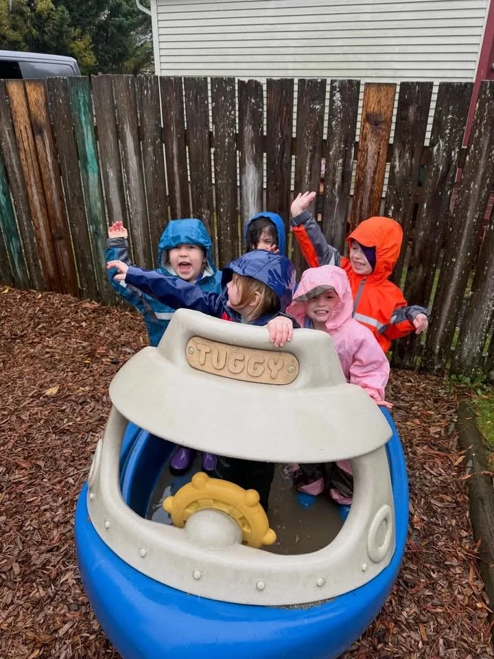 Four young children in colorful rain jackets and hats playing in a blue and white plastic tug boat in a backyard with a wooden fence and a house in the background.