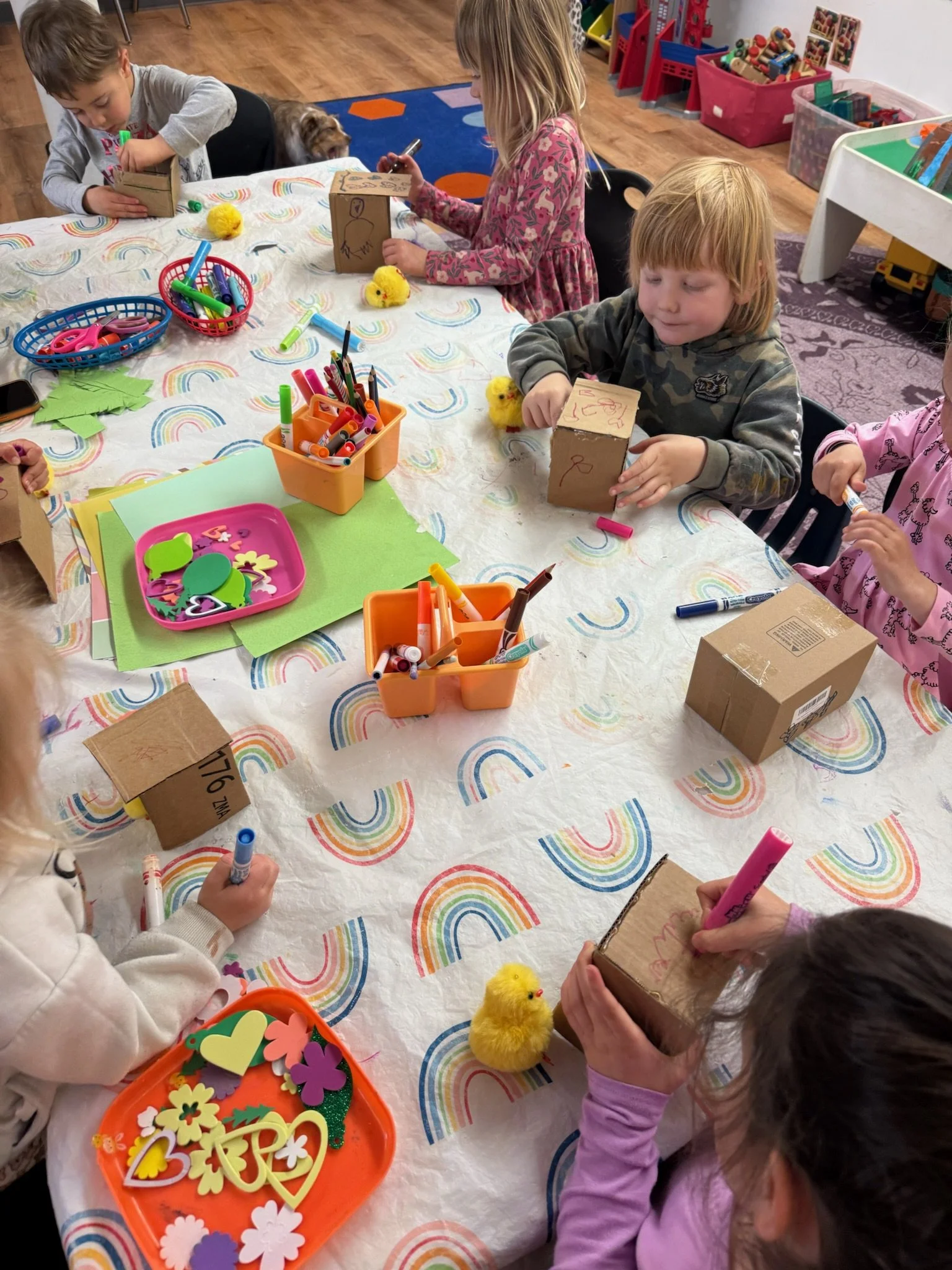 Children sitting at a table engaging in arts and crafts. The table has colorful markers, paper, and foam cutouts. A girl is holding a small cardboard box with drawings.