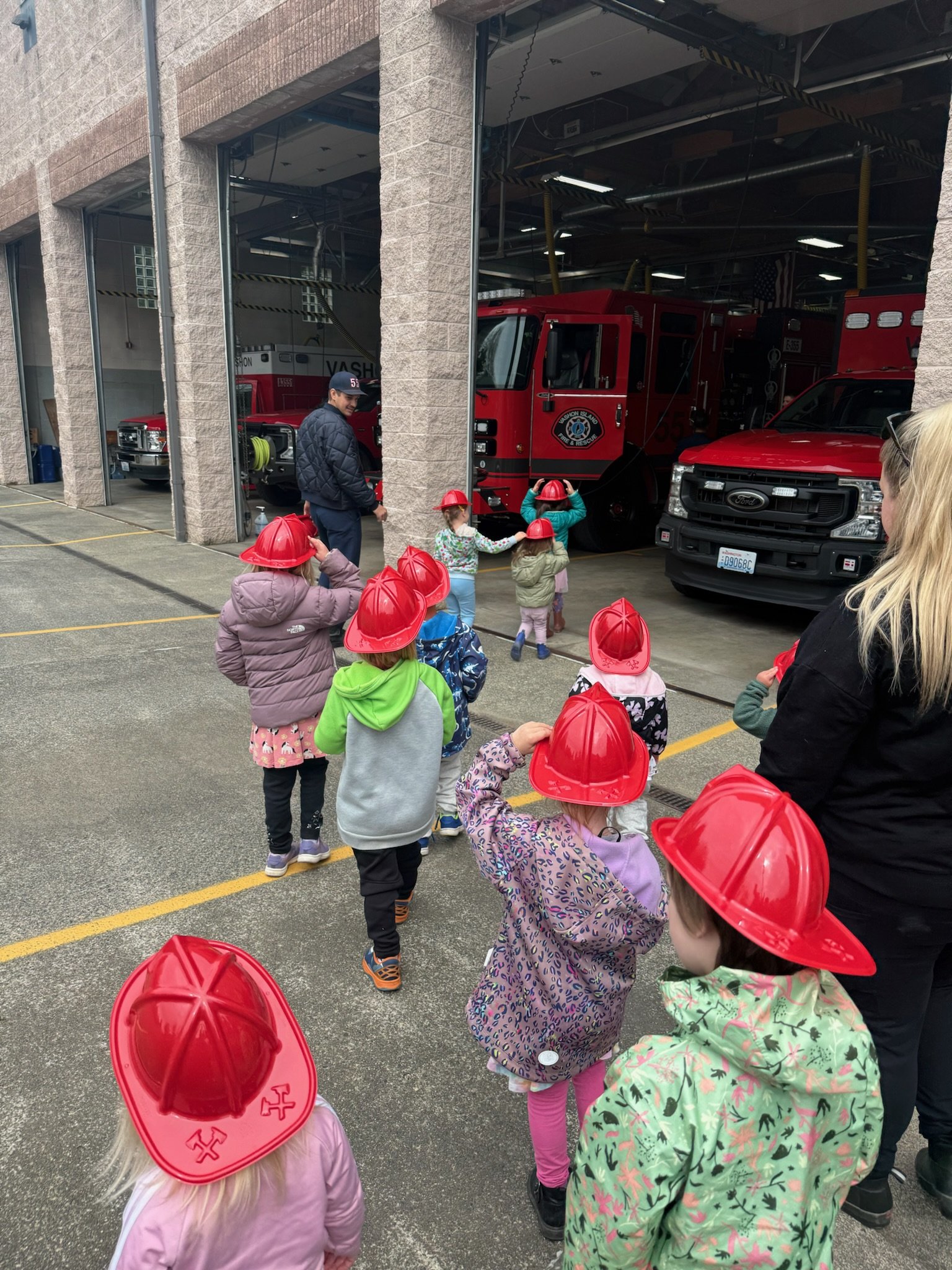 A group of children and adults with red fire hats, standing outside a fire station, observing fire trucks inside the station garage.
