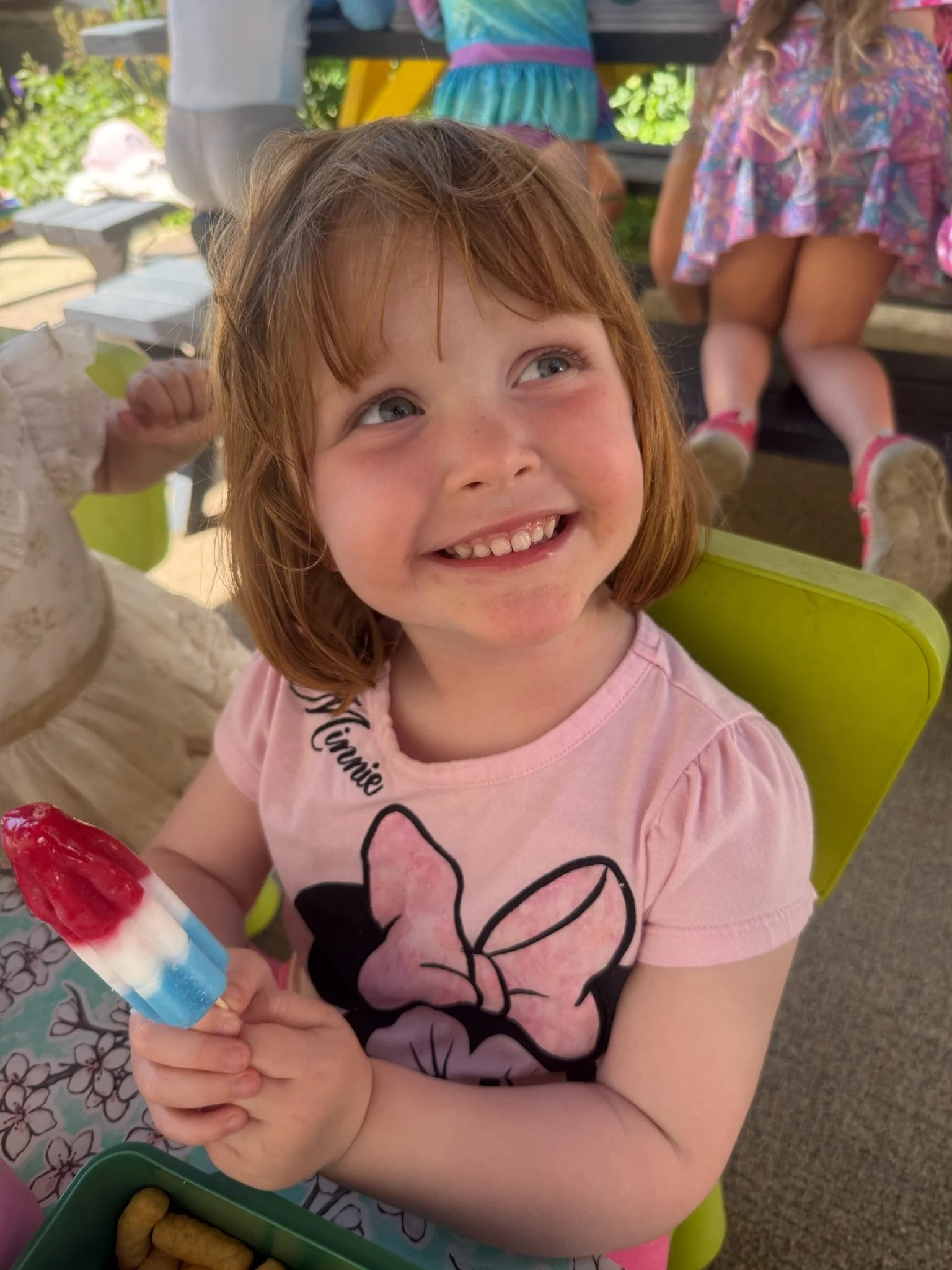 A young girl with red hair and a pink shirt featuring Minnie Mouse, smiling and holding a popsicle at an outdoor gathering. In the background, other children and some greenery are visible.