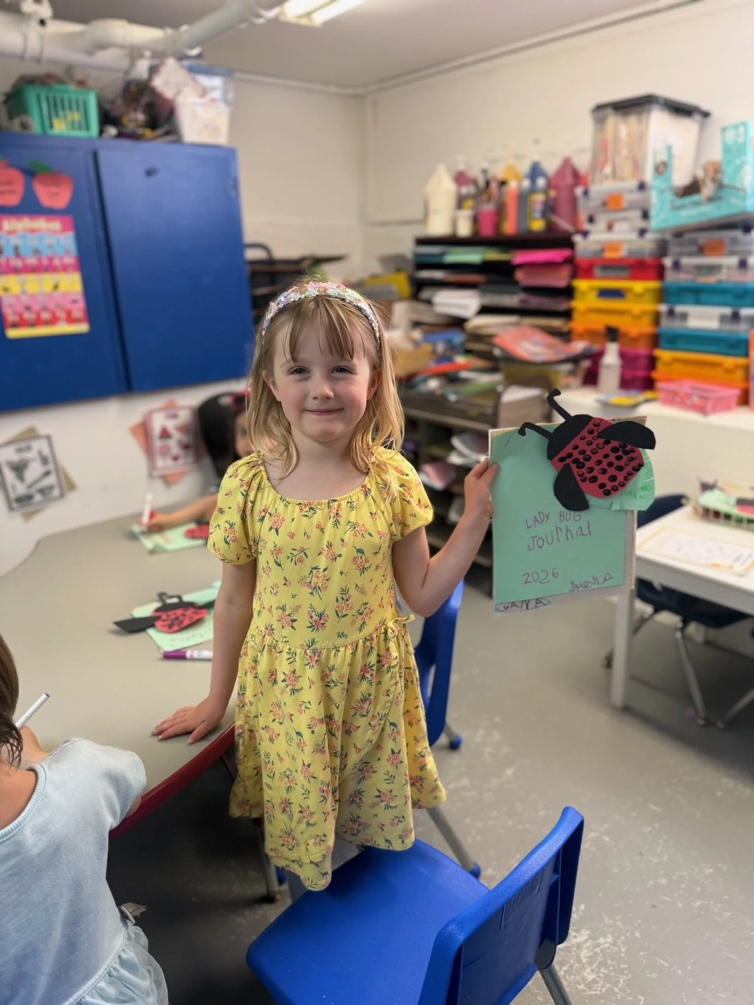 Young girl with blonde hair, wearing a yellow dress with pink floral pattern and a floral headband, holding a decorated ladybug craft project, standing in a classroom  with shelves filled with art supplies and classroom materials.