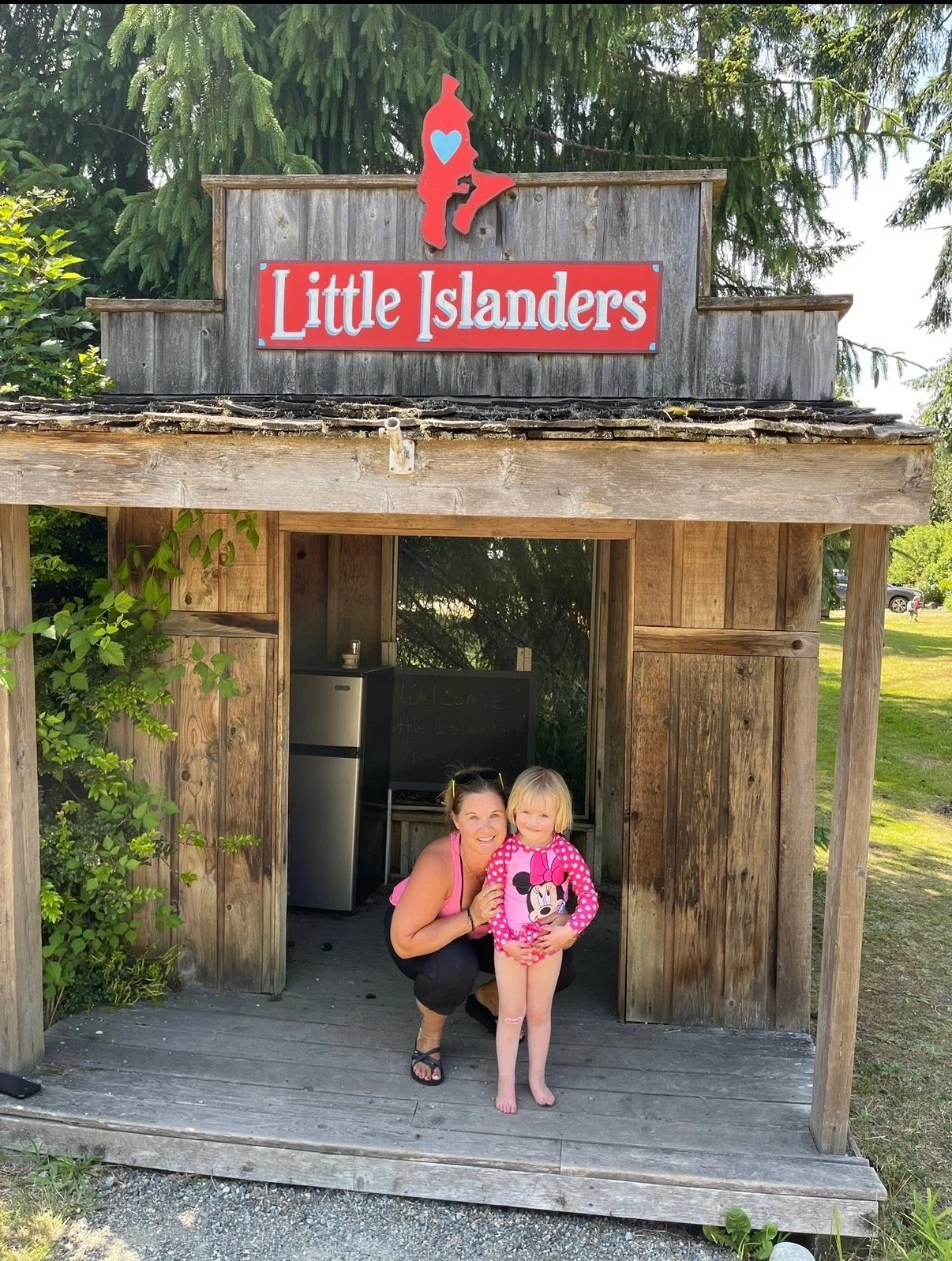 A woman and a young girl, both smiling, inside a small wooden playhouse labeled 'Little Islanders'. The girl is wearing a pink Minnie Mouse shirt and shorts, and the woman is wearing a pink tank top and black pants. The playhouse has a sign with a red shape and blue heart on top and is located outdoors with trees and grass around.