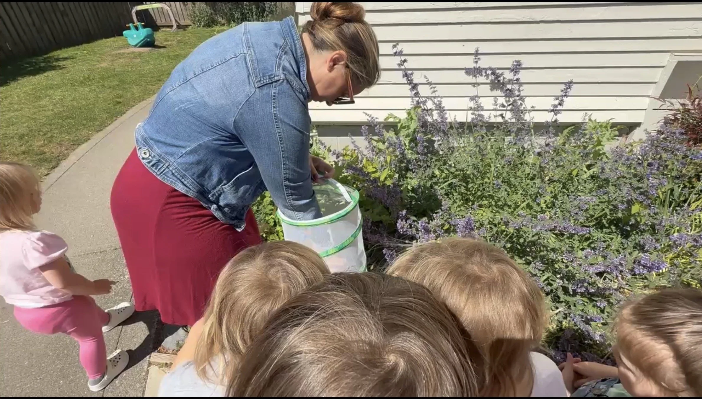 A woman with glasses and a denim jacket kneels by a garden bed with purple flowers, holding a white bucket, surrounded by young children with blonde hair, outside on a sunny day.