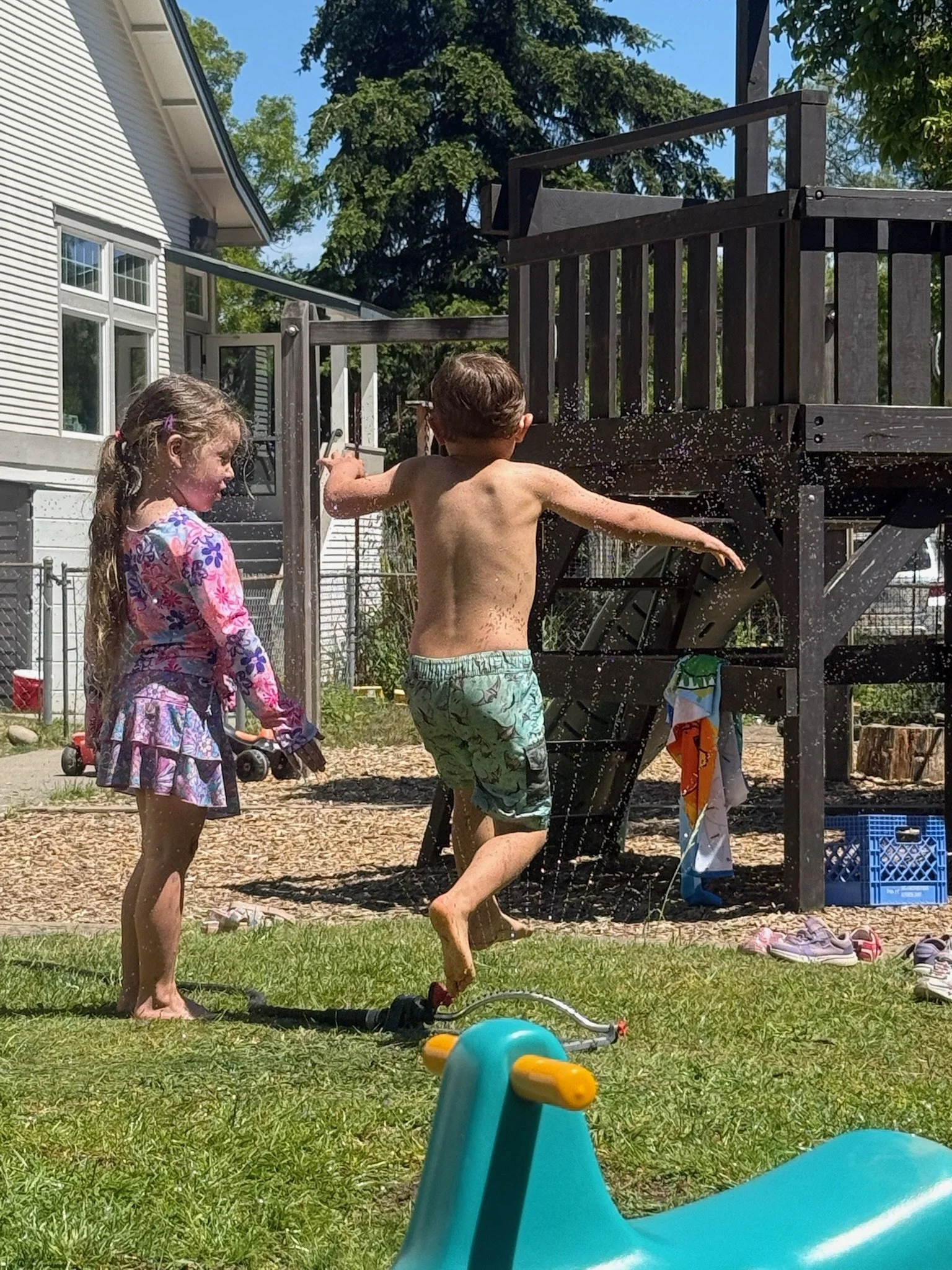 Two children playing in a backyard: a girl in a pink-and-purple dress holding a tricycle, and a boy in swim shorts jumping near a water sprinkler, with a playhouse slide and trees in the background.