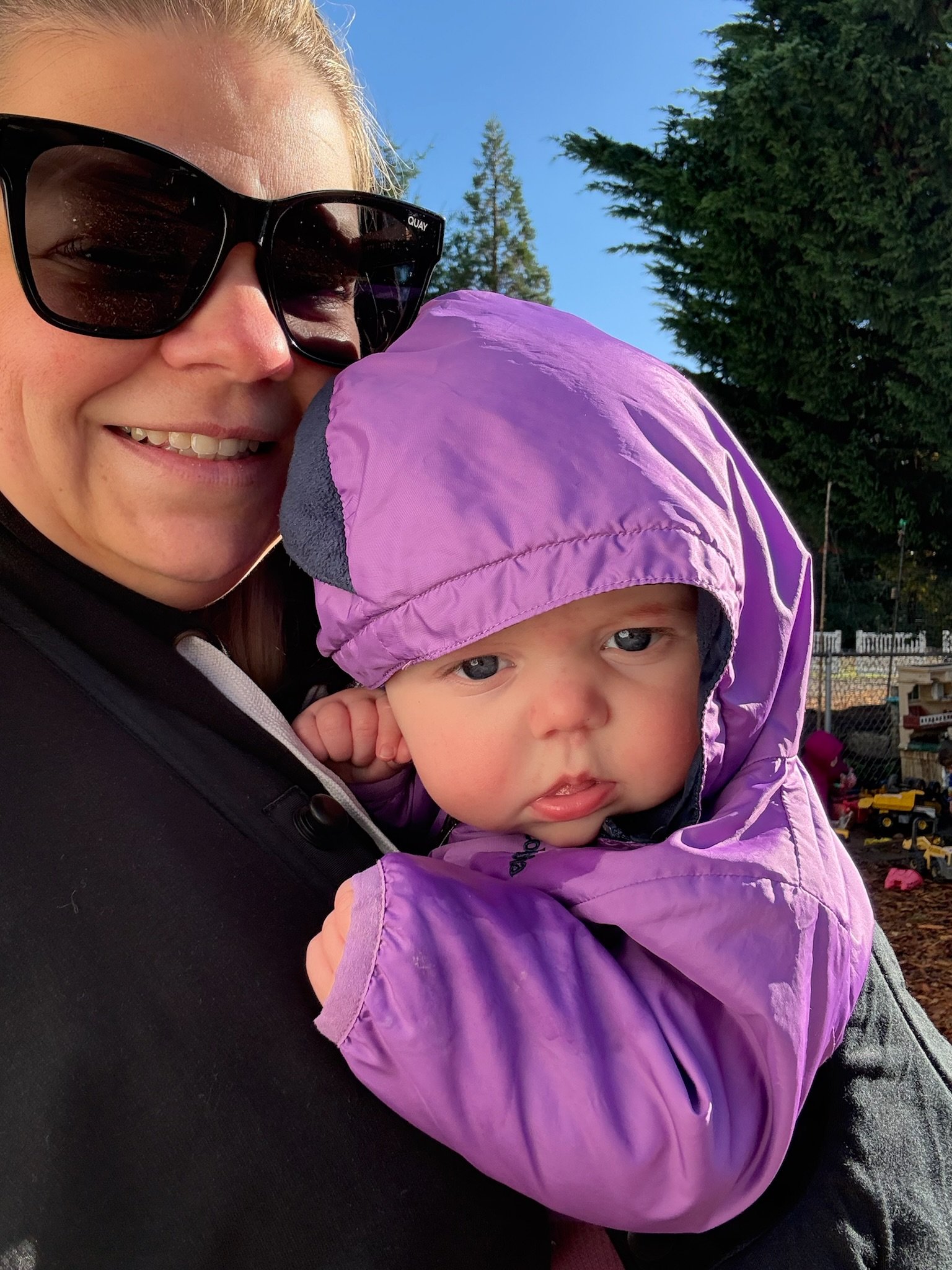 Close-up of a smiling woman wearing sunglasses holding a young child in a purple coat with a hood outdoors, with trees and a clear blue sky in the background.