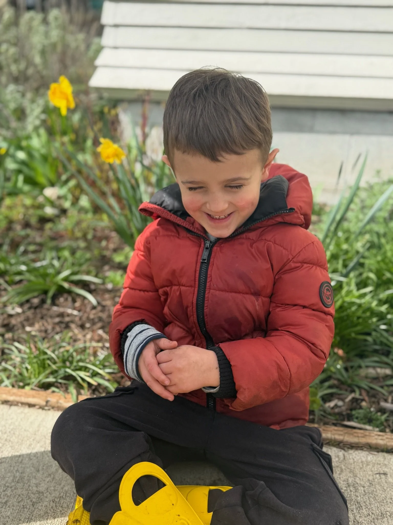 A young boy is sitting outdoors with closed eyes, smiling, wearing a red jacket, black pants, and striped gloves. He is sitting on the ground near yellow shoes and a black bag, surrounded by plants and yellow flowers.