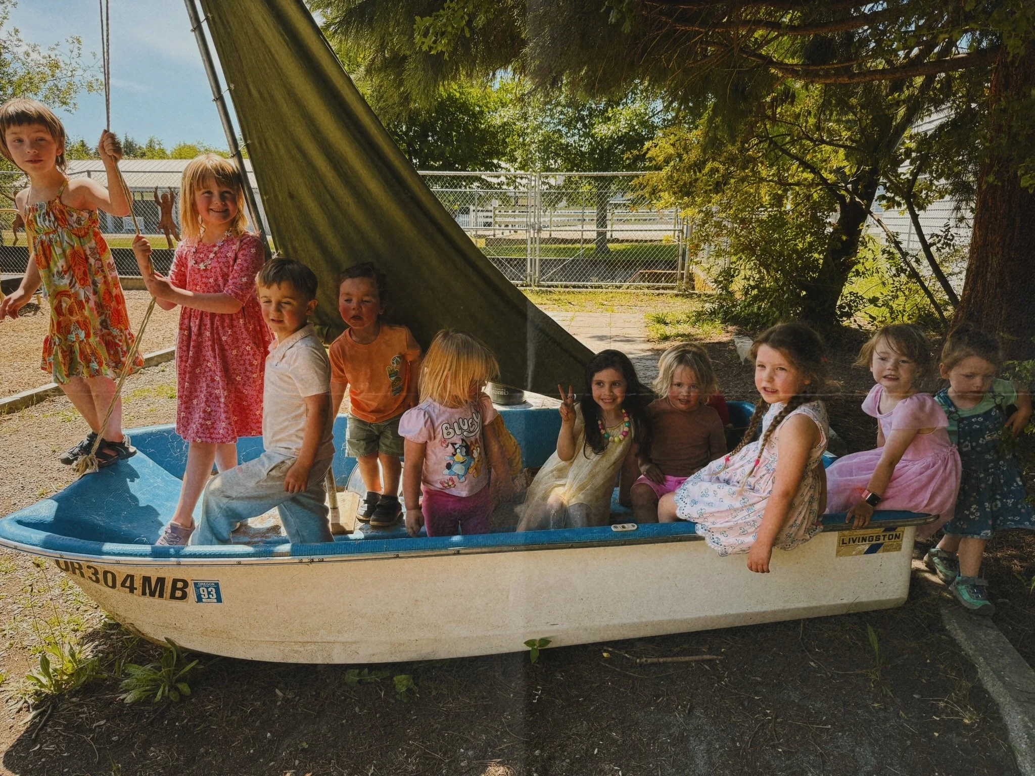 A group of children sitting and standing inside a small boat in an outdoor park setting with trees and a chain-link fence in the background.