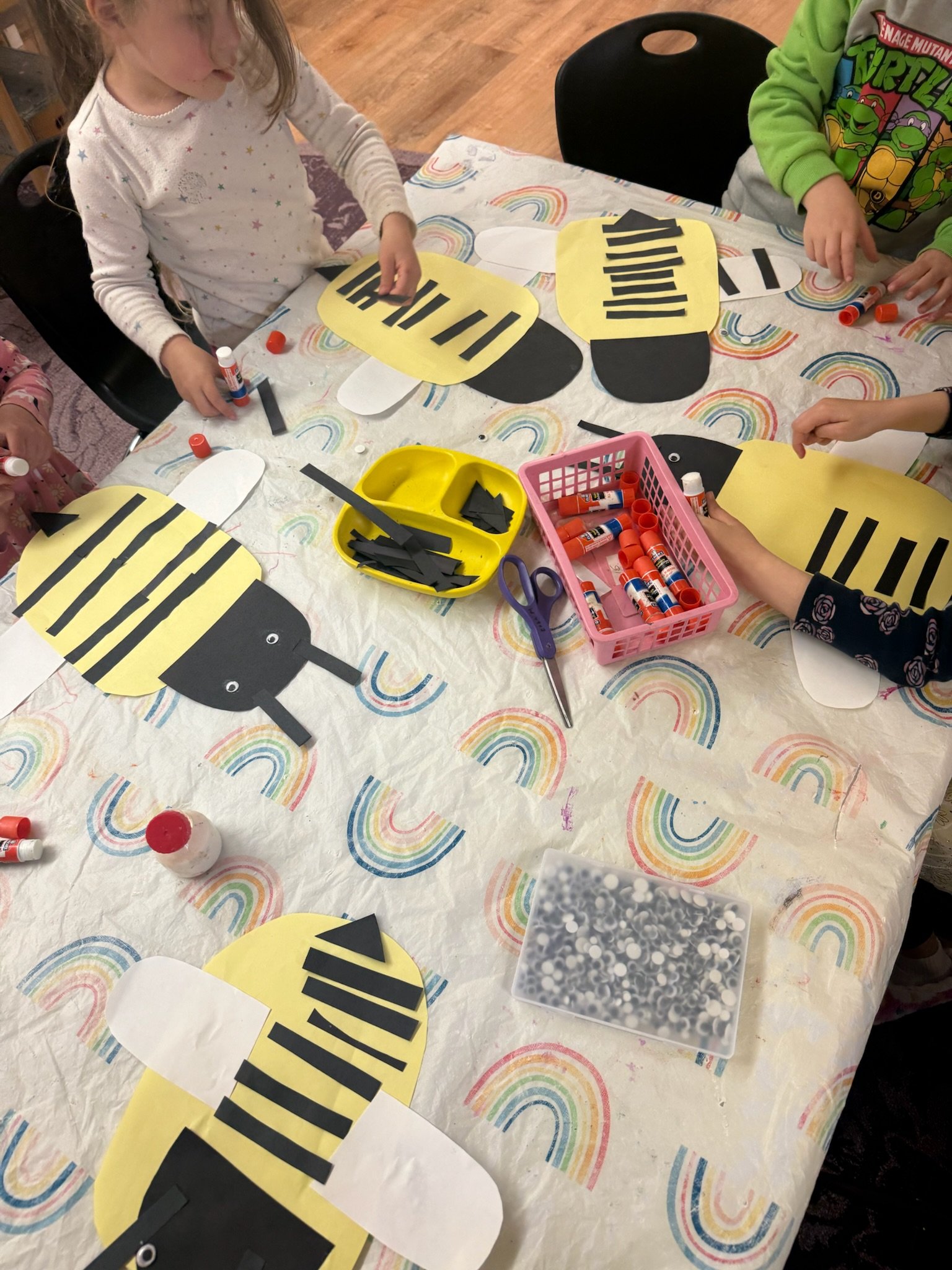 Children assemble black and yellow paper bees at a craft table covered with a rainbow rainbow pattern tablecloth.