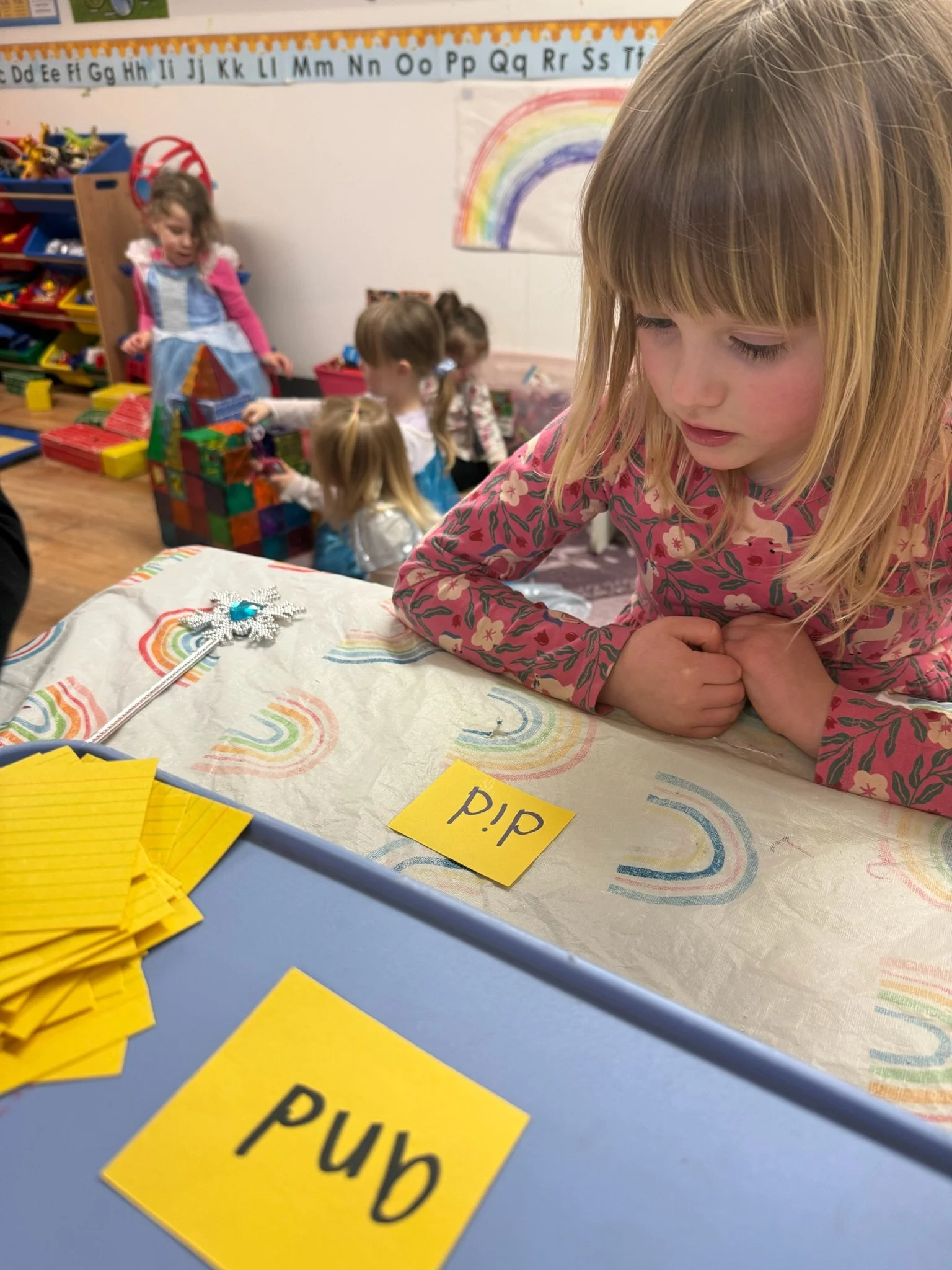 A young girl with blonde hair wearing a pink floral shirt laying on a table, with a colorful rainbow drawing in the background. In front of her, there are yellow cards and several yellow paper strips.
