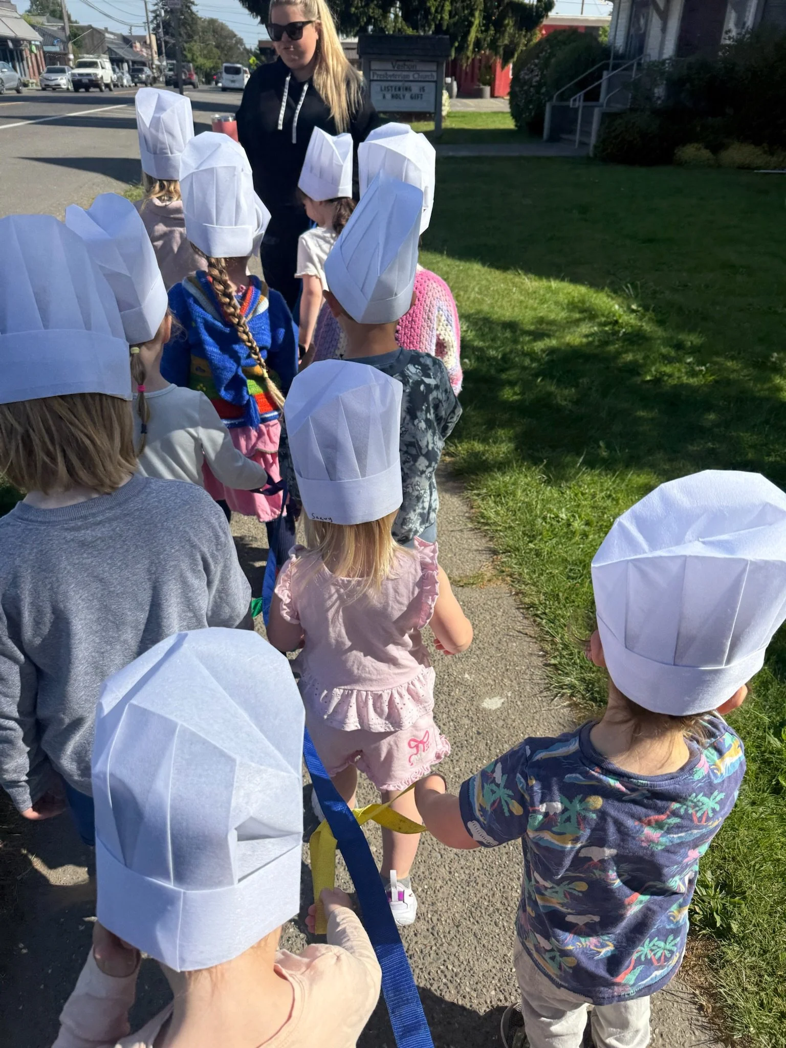 A group of children wearing white chef hats walking outdoors on a sidewalk, accompanied by an adult woman with blonde hair and sunglasses. The children are holding hands, and the setting appears to be a neighborhood or small town with houses, trees, cars, and a street in the background.