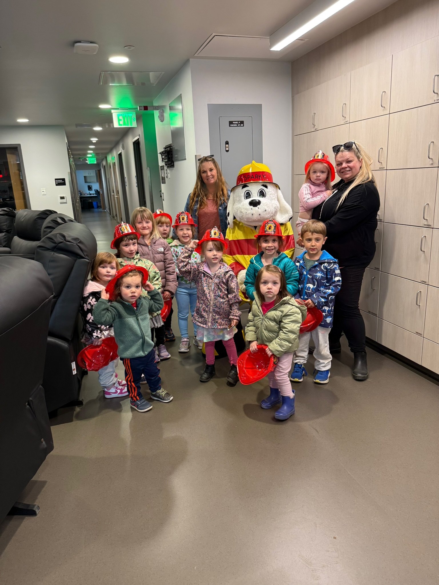 A group of young children and two adult women gathered in a hallway, dressed casually, some wearing red firefighter helmets, in front of a person in a large dog mascot costume dressed as a firefighter, with lockers and exit signs visible in the background.