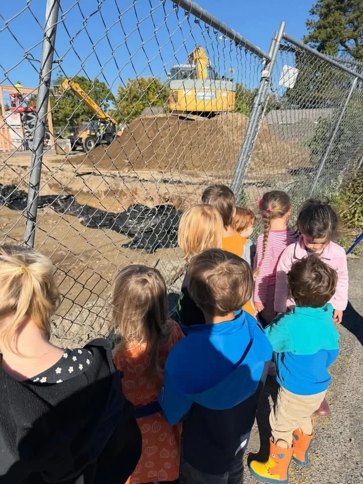 Children observing a construction site with a yellow excavator through a chain-link fence.