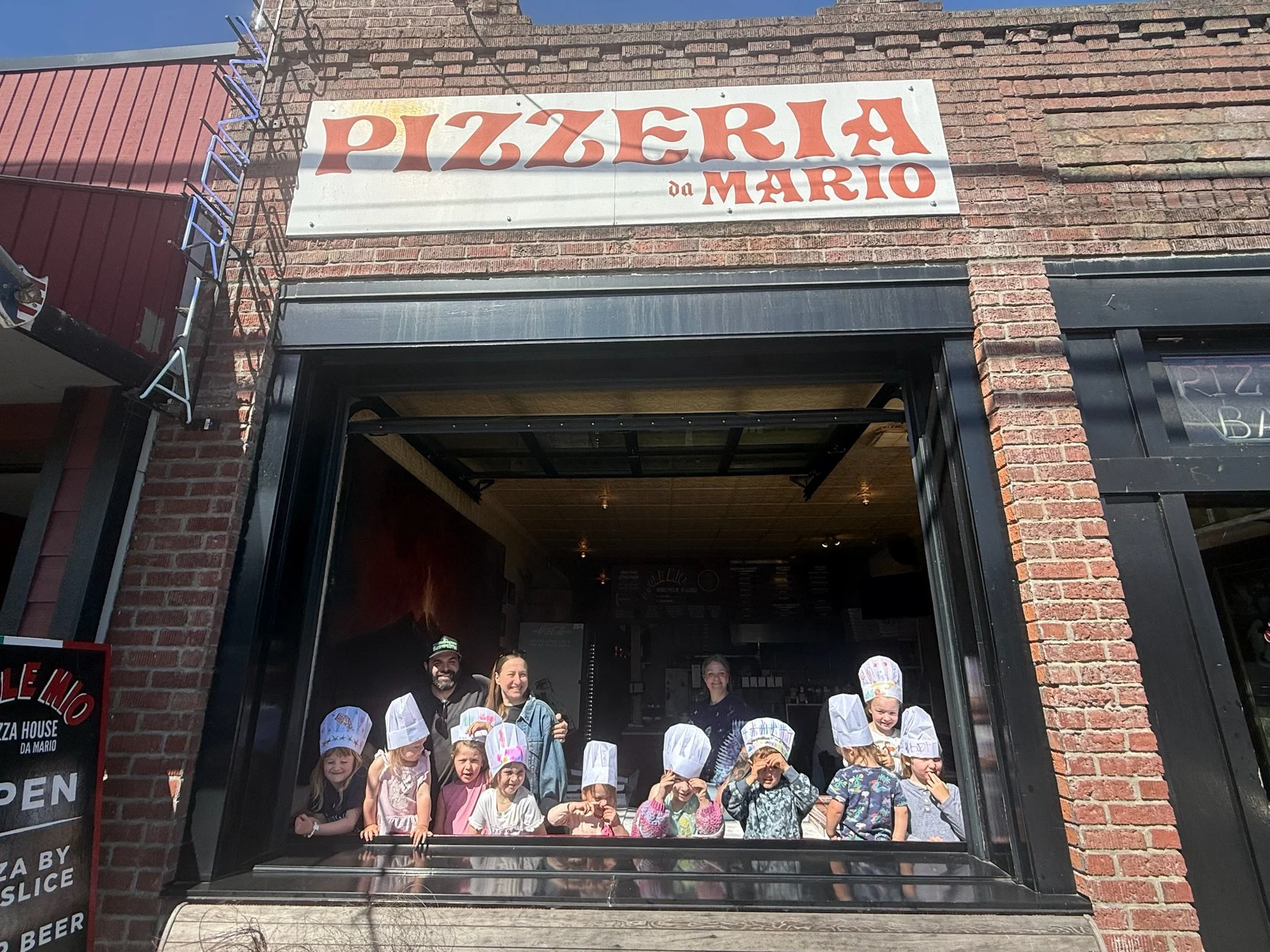Children and adults standing together outside a brick pizza restaurant with a sign that reads "Pizzeria da Mario." The children are wearing paper chef hats, and the group appears to be posing for a photo.