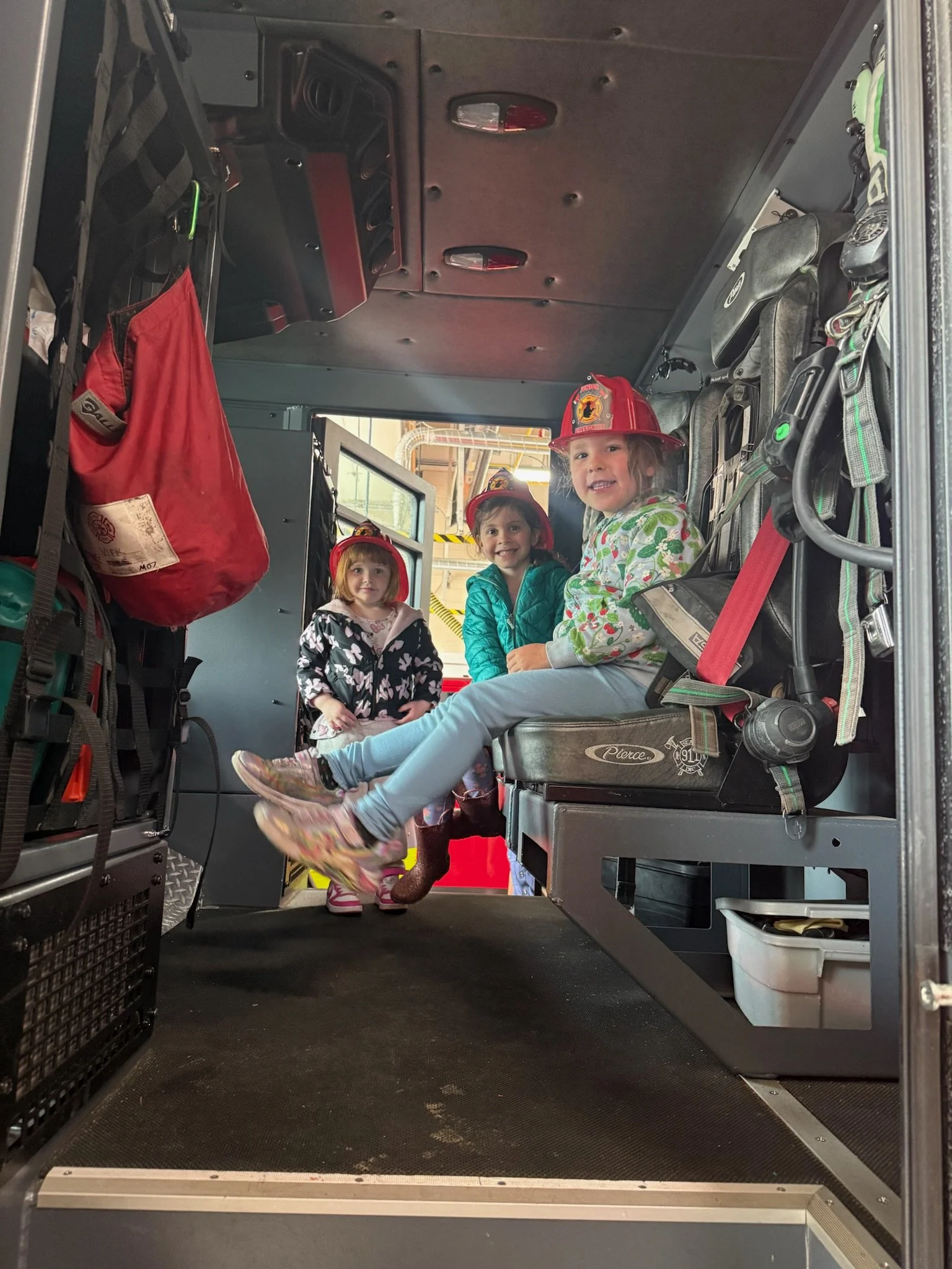 Three young girls in firefighter helmets inside a fire truck at a fire station, smiling at the camera.