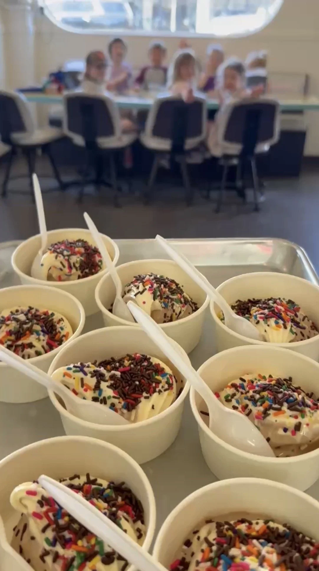 Bowls of vanilla ice cream topped with colorful sprinkles and chocolate sprinkles, with white plastic spoons, in a kitchen or cafeteria setting.