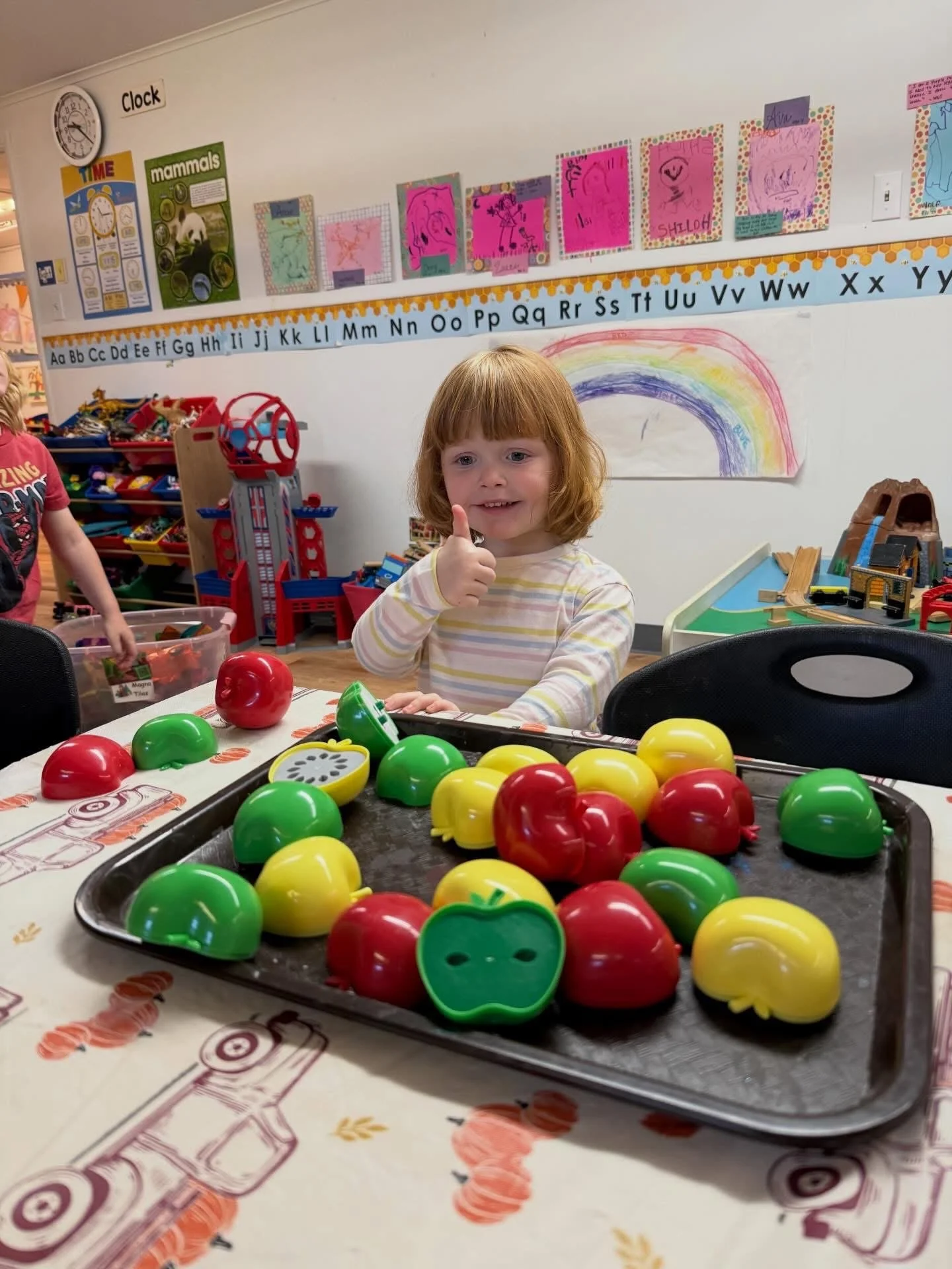 Young girl with red hair smiling and giving a thumbs up at a table with colorful plastic apples and peppers, in a classroom decorated with educational posters and children's artwork.