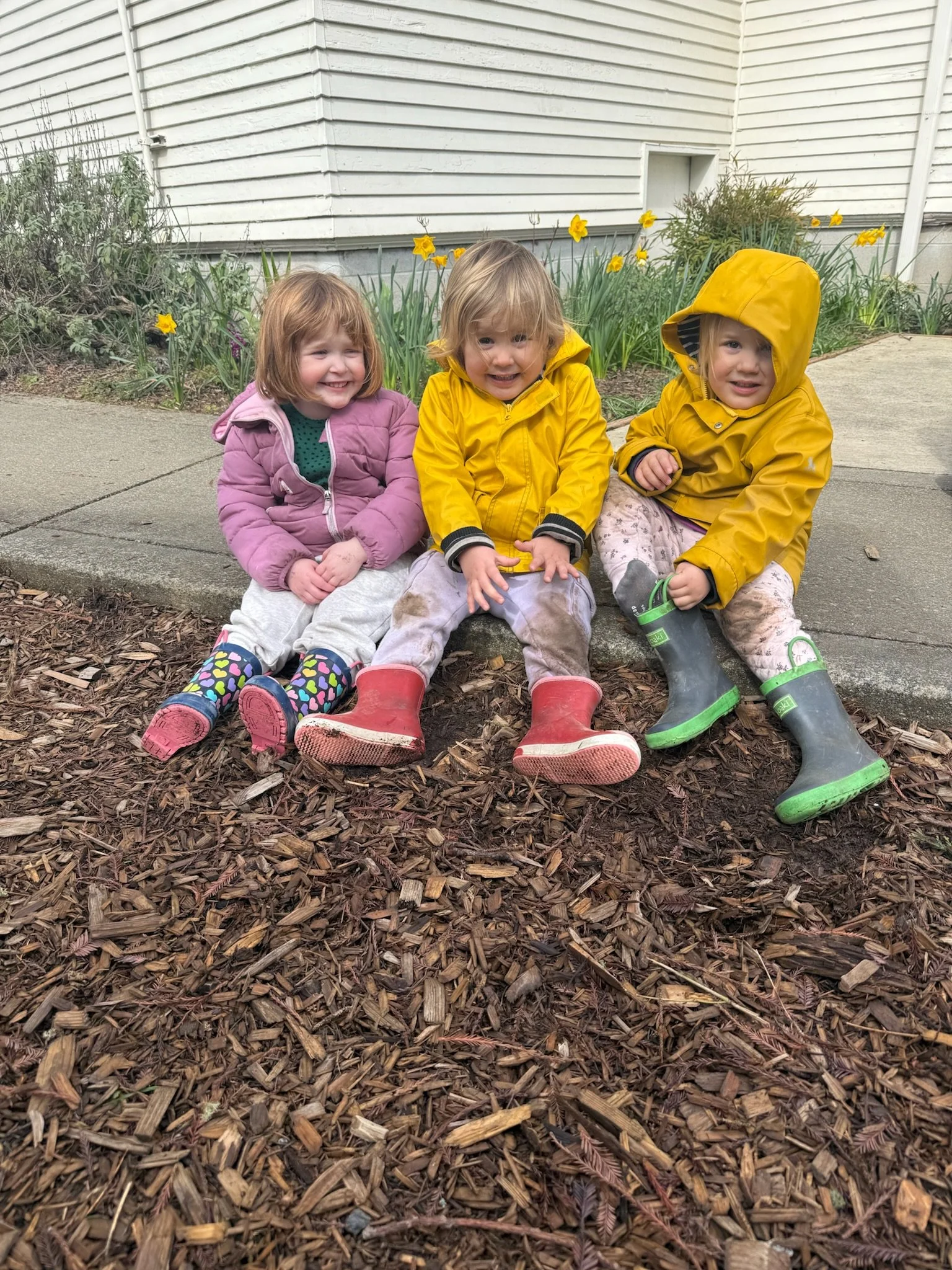 Three young children sitting on a mulch-covered ground in front of a flower bed with yellow flowers, in front of a white house with horizontal siding.