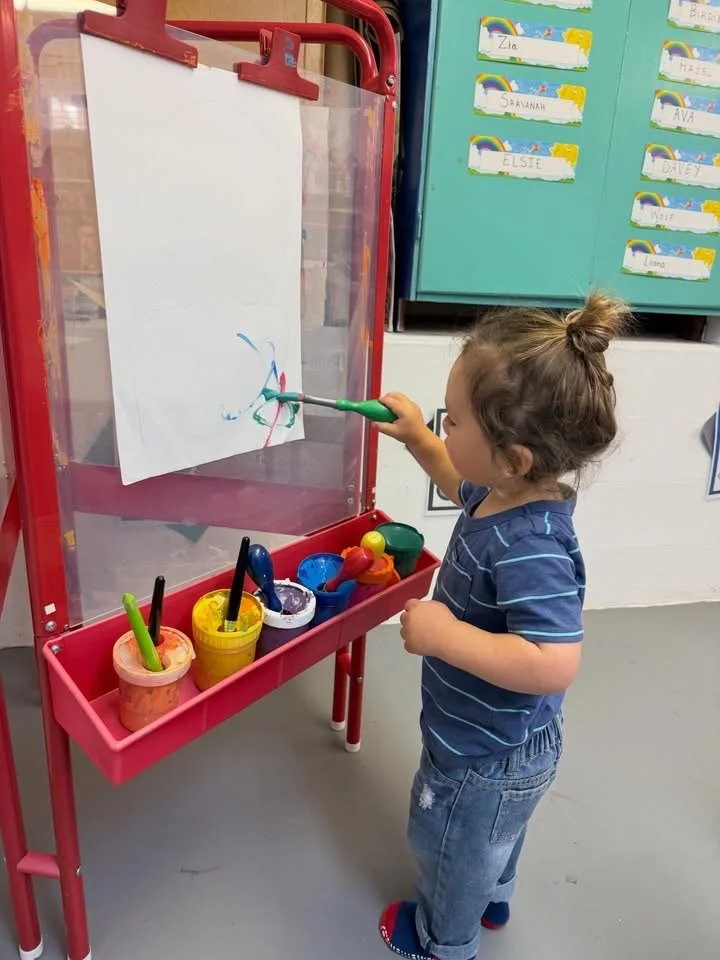 A young girl with brown hair tied in a bun is painting with green and blue paint on a clear plastic surface with a green marker. The girl wears a blue striped shirt and jeans, and stands in front of a red paint easel with multiple cups of colorful paints nearby. Behind her are colorful lockers with name tags.