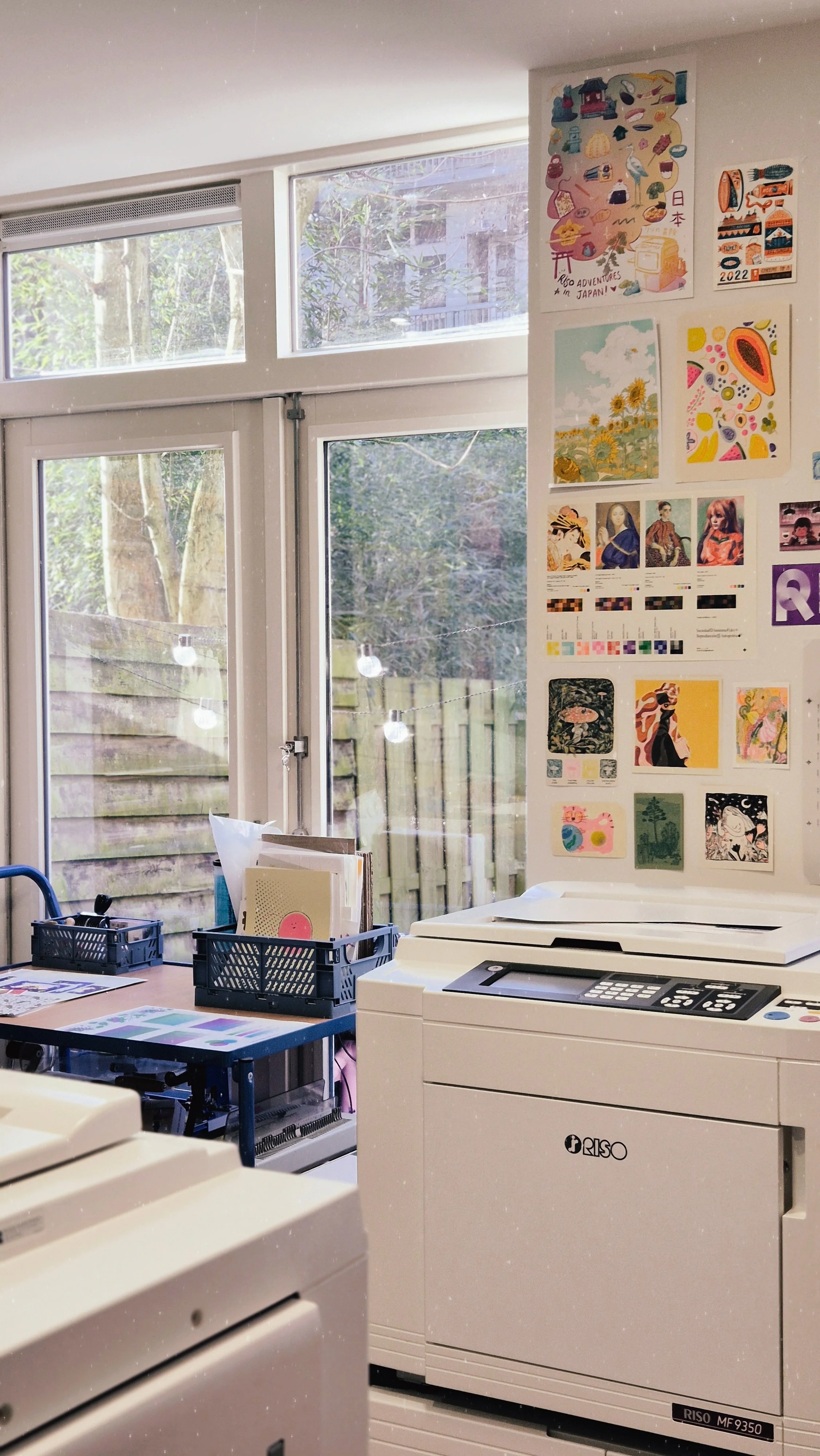 Photo of a studio with riso printer visible in the foreground. There are some riso prints on the wall and on an adjacent table.