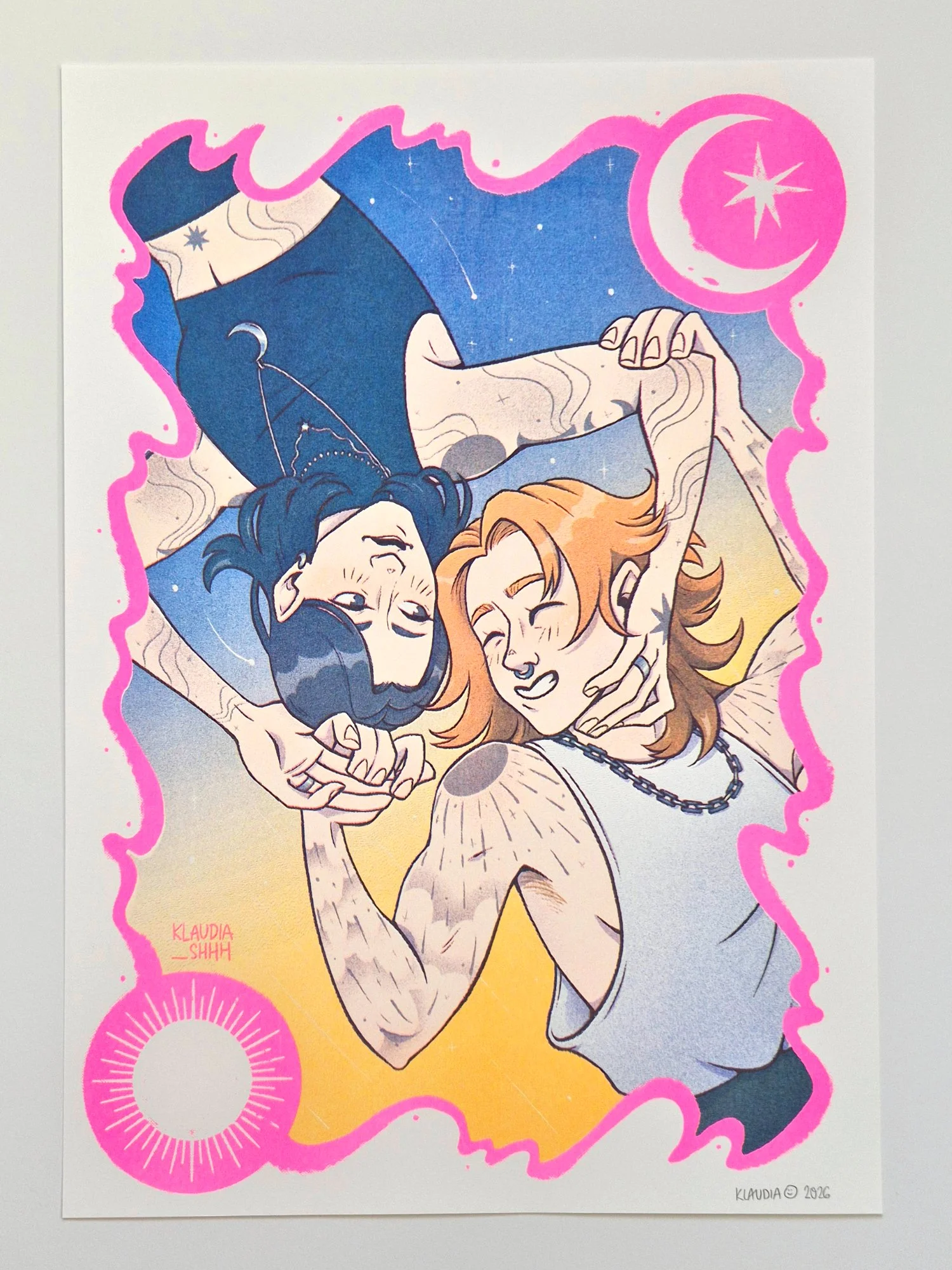 A photo of an A3 risograph print showing two women floating through the air, embracing. One represents the moon, while the other is the sun. The sky behind them has twilight colours.