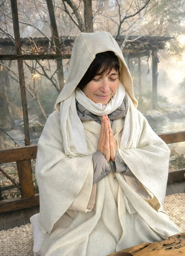 A woman dressed in white robes with a hood, praying with hands in a prayer position, sitting outdoors near a body of water and trees.