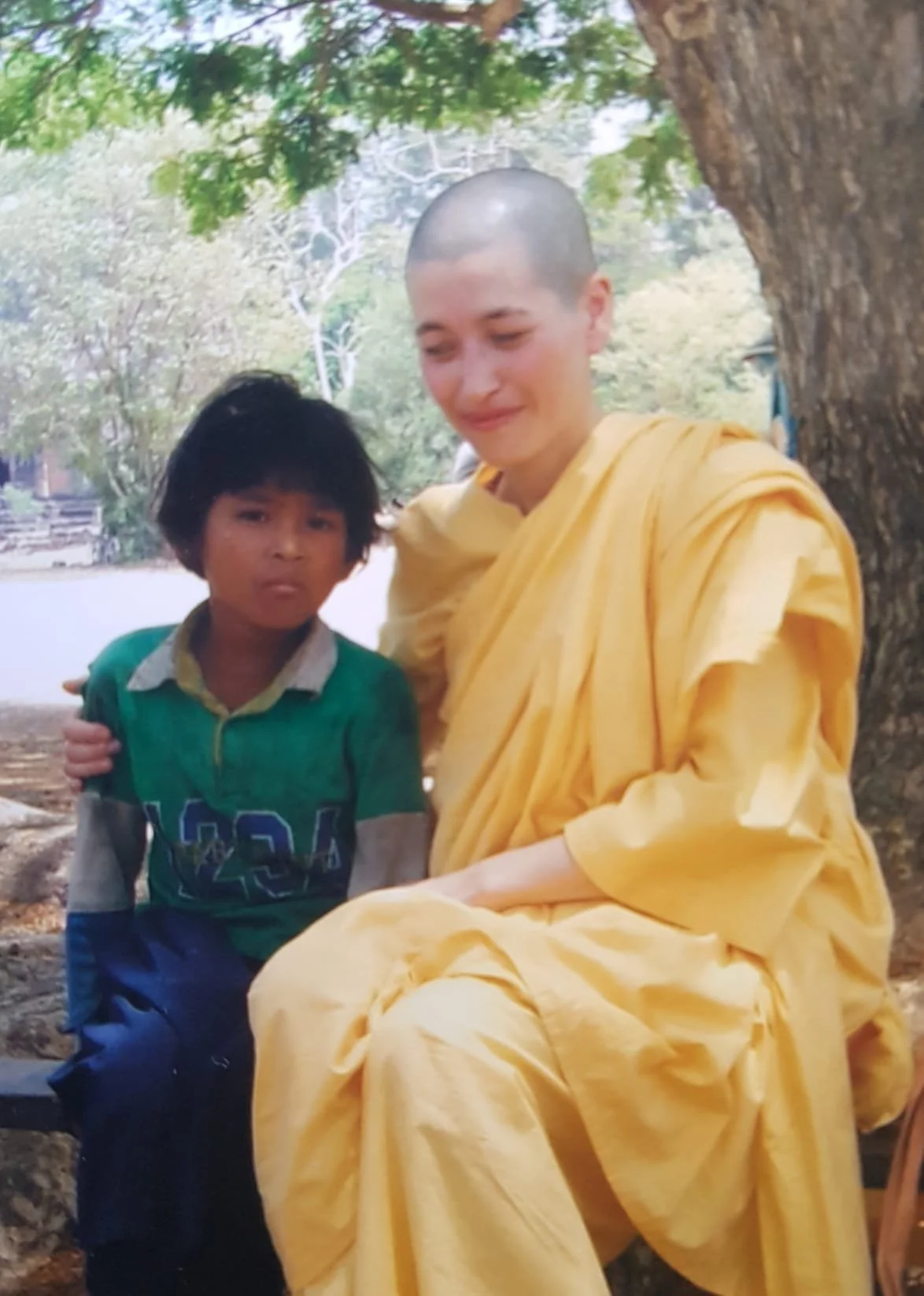 A young boy with dark hair, wearing a green and gray shirt, sitting next to a woman with a shaved head, wearing a yellow robe, outdoors near a tree with green leaves.