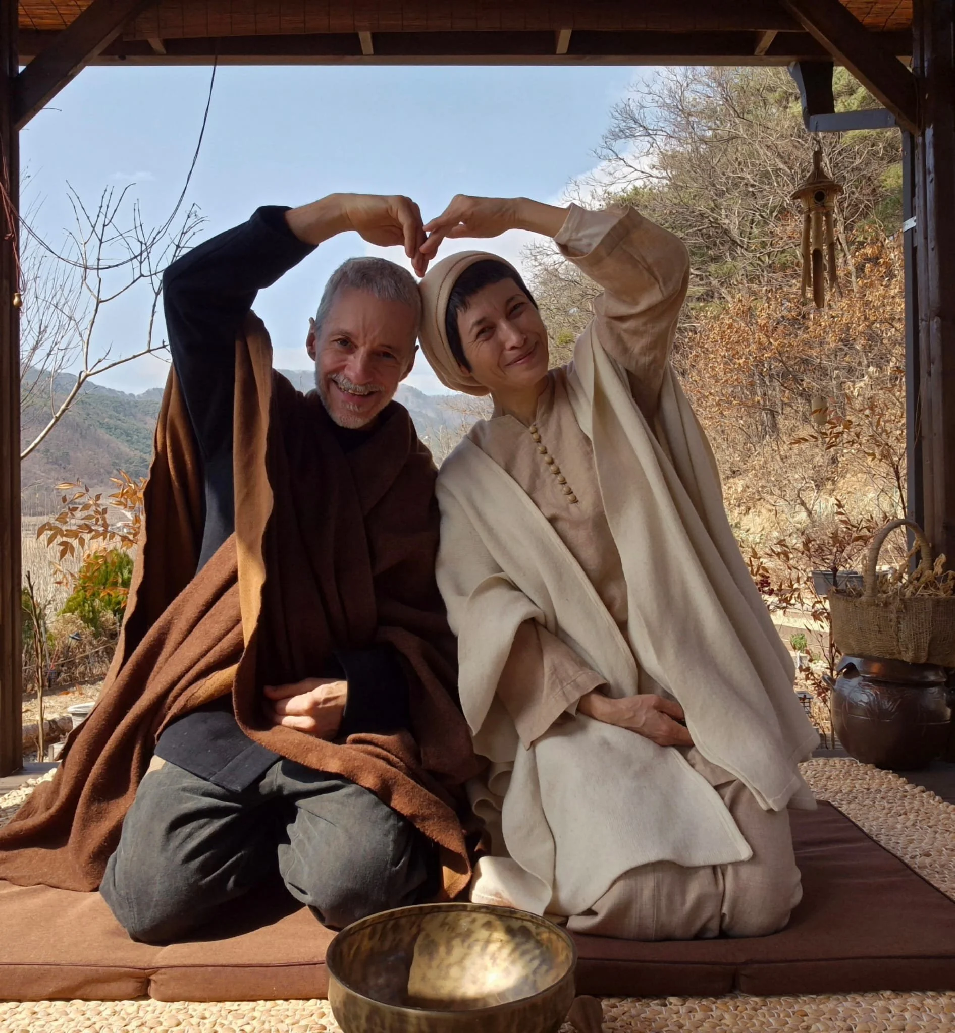 A smiling man and woman are sitting on a cushion on the floor inside a wooden structure, making a heart shape with their hands. The background shows a scenic outdoor landscape with hills and trees, some with autumn-colored leaves. There are decorative items like a wind chime and a basket nearby.