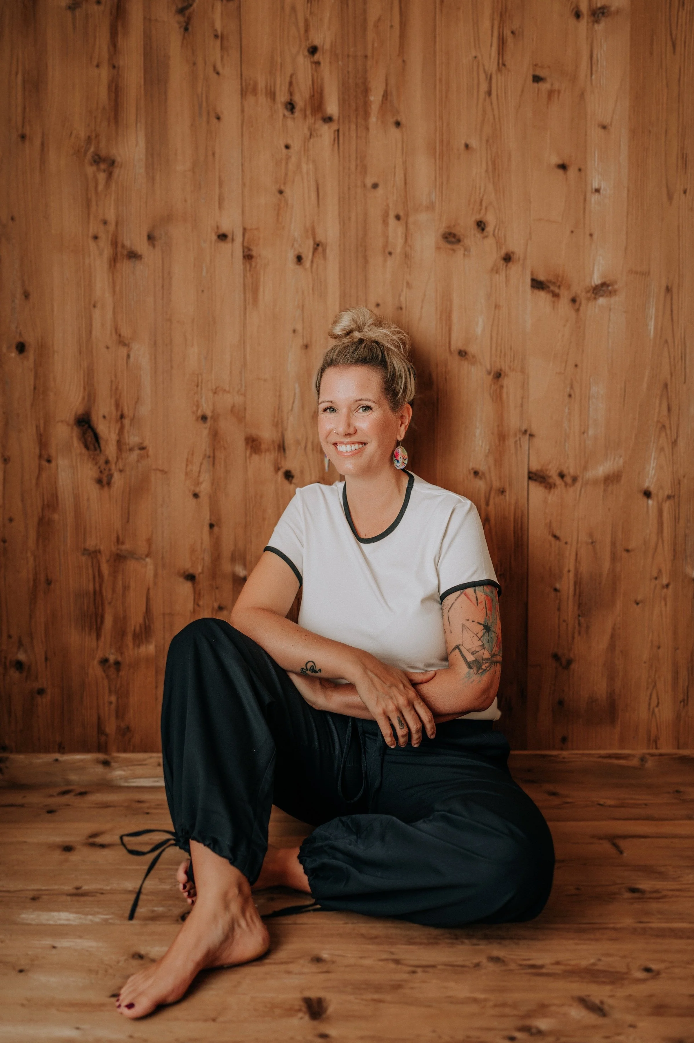 Junge Frau mit Tattoos sitzt auf dem Holzboden vor einer Holzvertäfelung, lächelt und trägt weißes T-Shirt und schwarze Hosen.