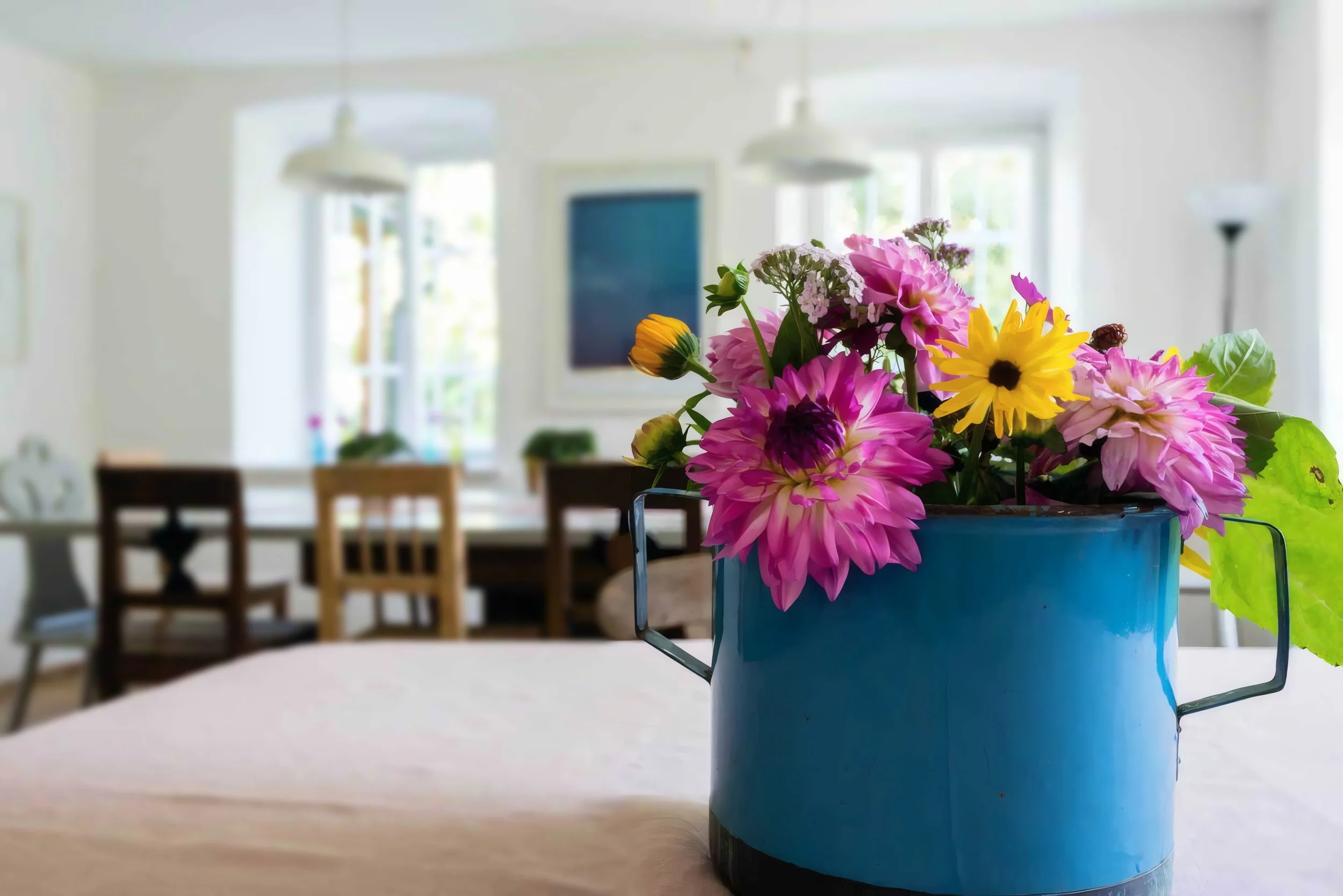 Blumen in einer blauen Dose auf einem Tisch in einem hellen Raum mit großen Fenstern und mehreren Stühlen im Hintergrund.